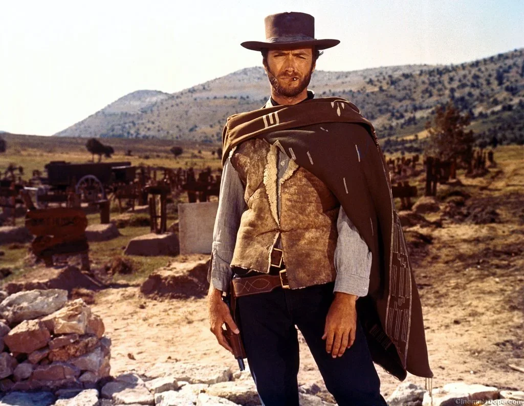 A man dressed as a cowboy standing in a historic Western cemetery with graves and mountains in the background.