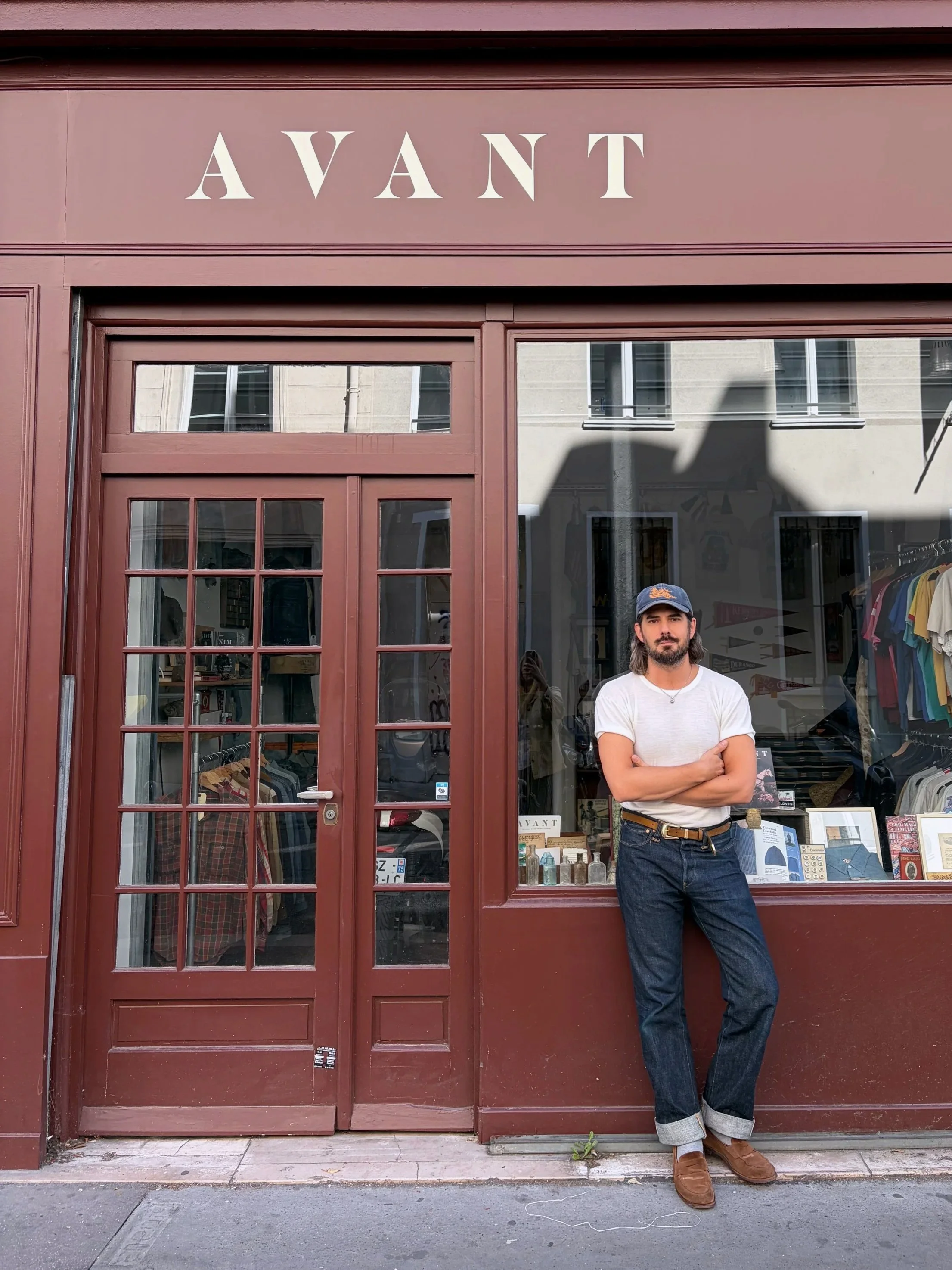 A man with dark hair and a beard wearing a light-colored wide-brimmed hat, a cream-colored shirt with brown patterned embroidery, and crossed arms standing in a clothing store.