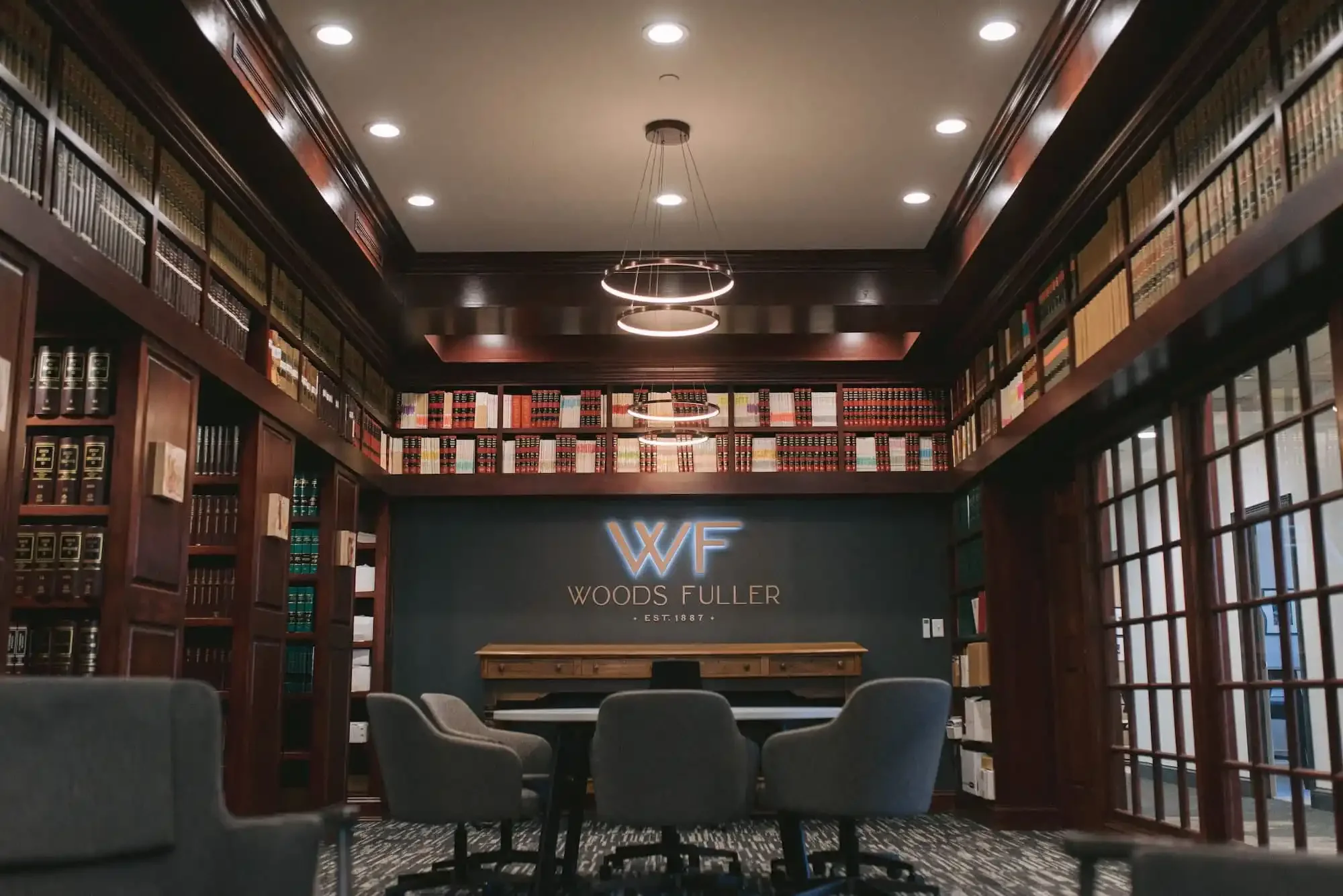 A modern library conference room with dark wood bookshelves filled with books, a central table with four gray chairs, and a sign reading 'WF Woods Fuller EST. 1887' on a dark wall, with circular ceiling lights and large windows on the right side.