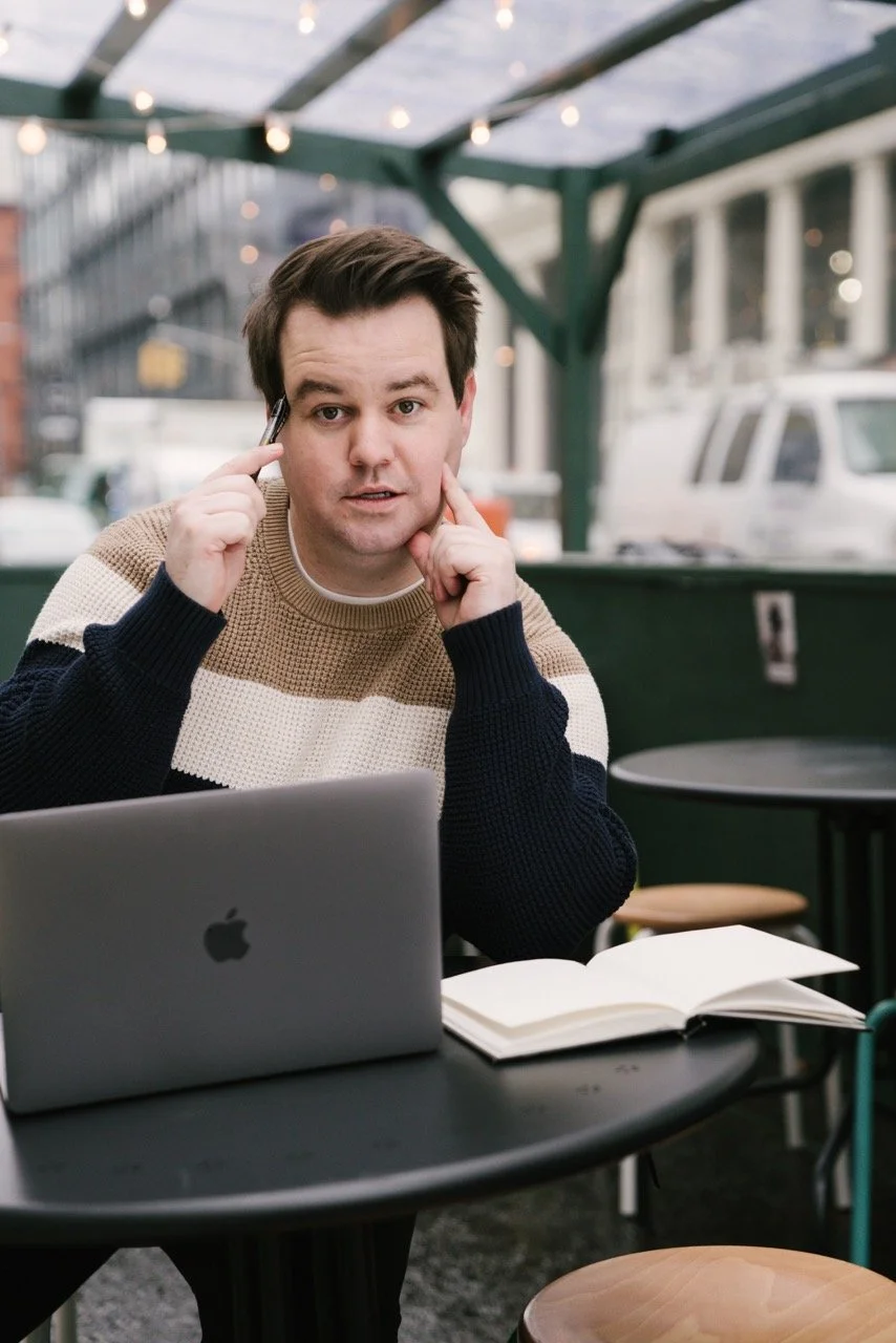 A young man sitting at an outdoor cafe table with a silver MacBook, open book, and smartphone, wearing a beige, white, and navy sweater, with city buildings and parked cars in the background.