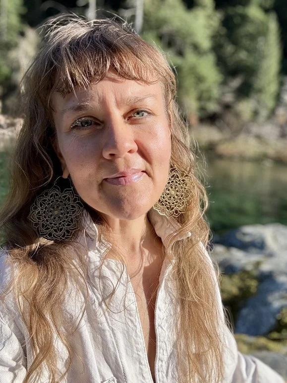 A woman with long light brown wavy hair, light skin, and blue eyes standing outdoors near a river with rocks and trees in the background. She is wearing large, intricate earrings and a white shirt, looking at the camera with a slight smile.