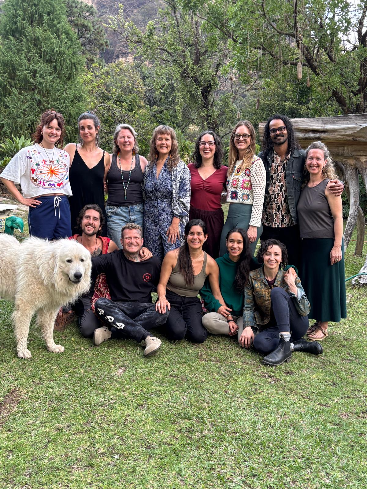 A group of fifteen people, mostly women, and a large white dog, posing outdoors in a green, wooded area with trees and rocks in the background.