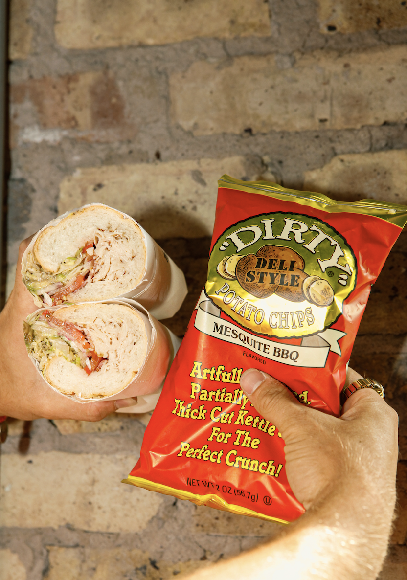 Person holding a bag of Dirt Potato Chips Mesquite BBQ flavor and a sandwich wrap with lettuce, tomato, and turkey in front of a brick wall.