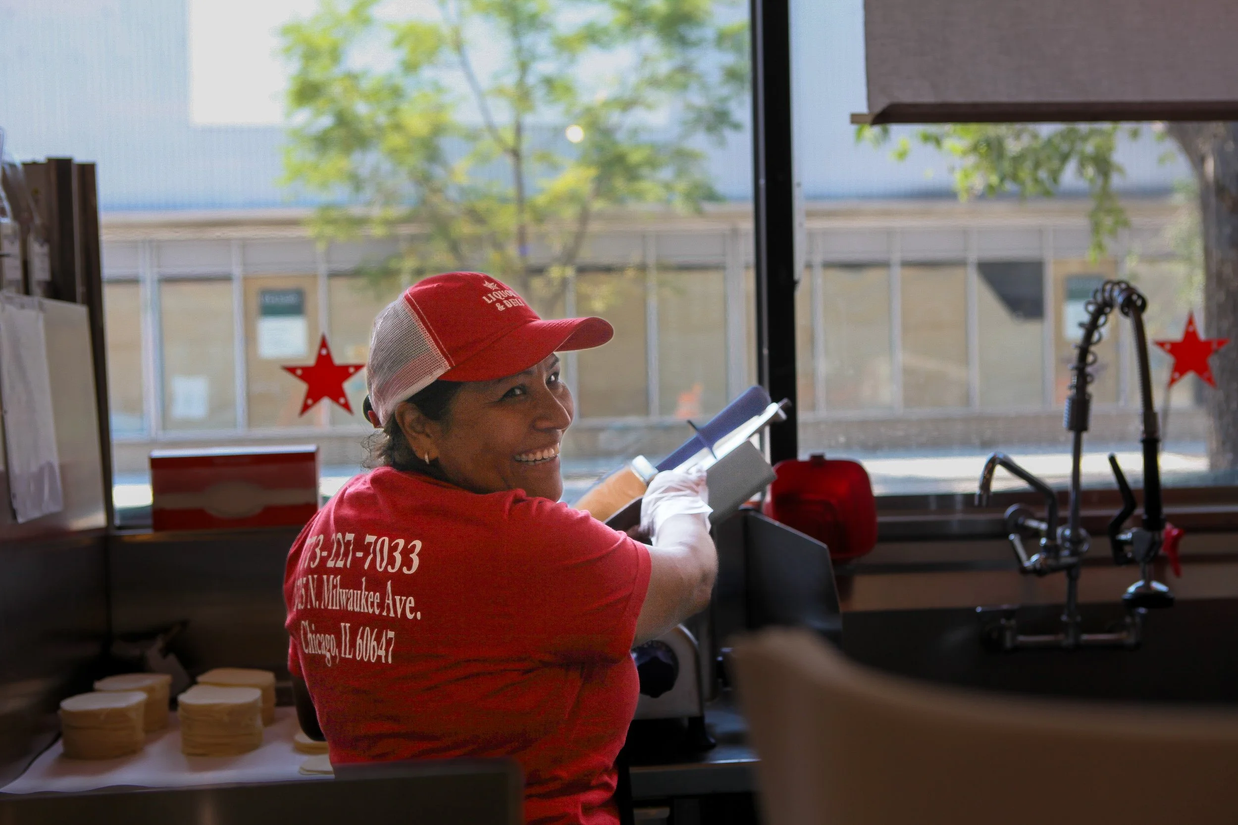 A woman wearing a red cap and red uniform working behind the counter at a food establishment, with green trees visible outside the window.