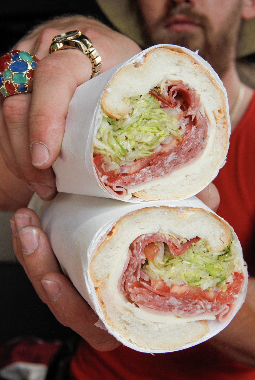 Person holding two large deli wrap sandwiches filled with lettuce, tomato, and various deli meats.