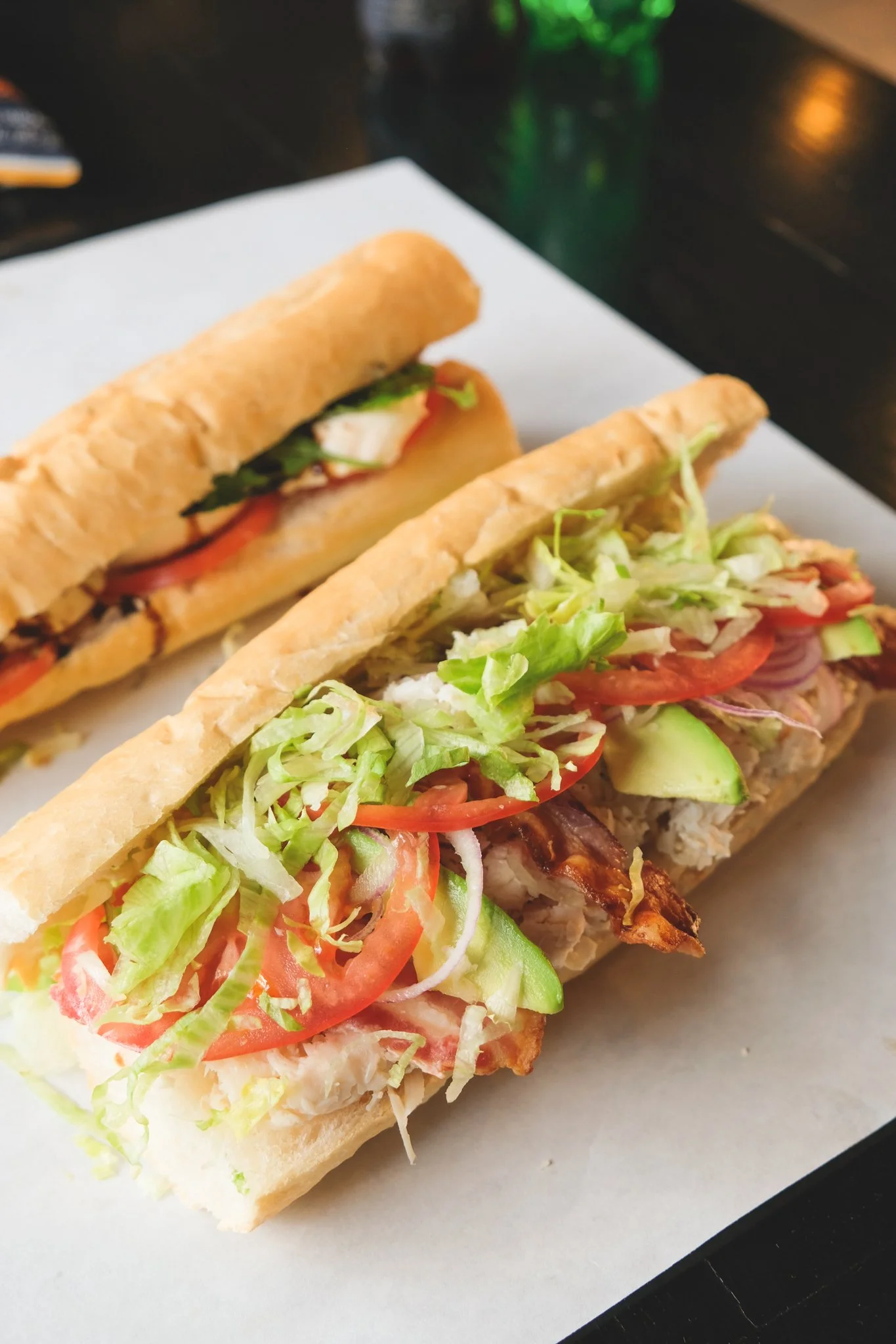Close-up of two subs with fresh vegetables on a white sheet of paper, with green and brown beverages in the background.