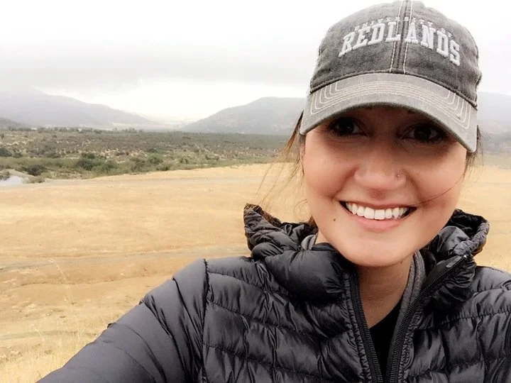 Close-up of a smiling woman wearing a gray cap and black puffy jacket outdoors with a mountains and cloudy sky background.