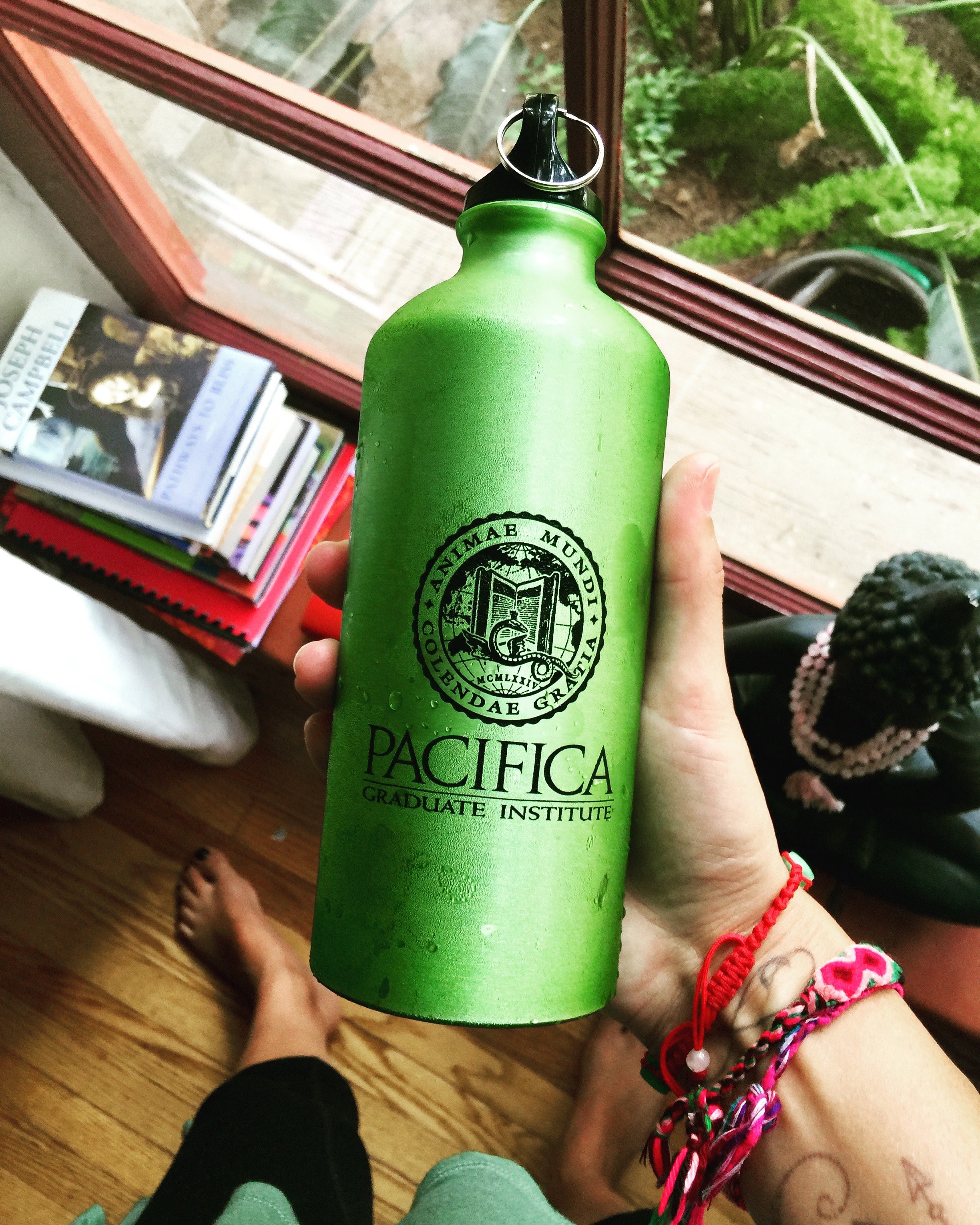 A hand holding a green water bottle with the logo of Pacifica Graduate Institute, featuring an open book and a serpent, near a window with a view of plants outside. There are books and a potted plant nearby, and the person's feet are visible on a wooden floor.