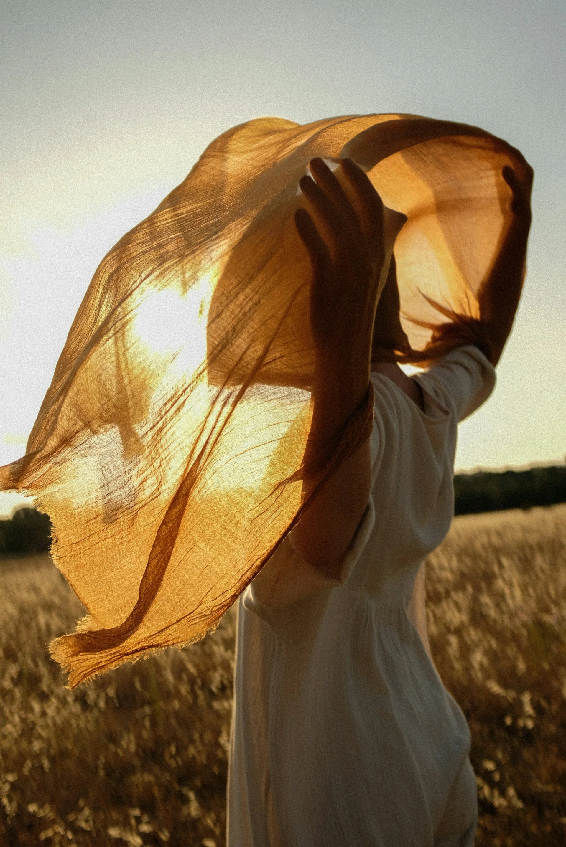 Person holding a light-colored cloth or scarf over their head in a field during sunset.
