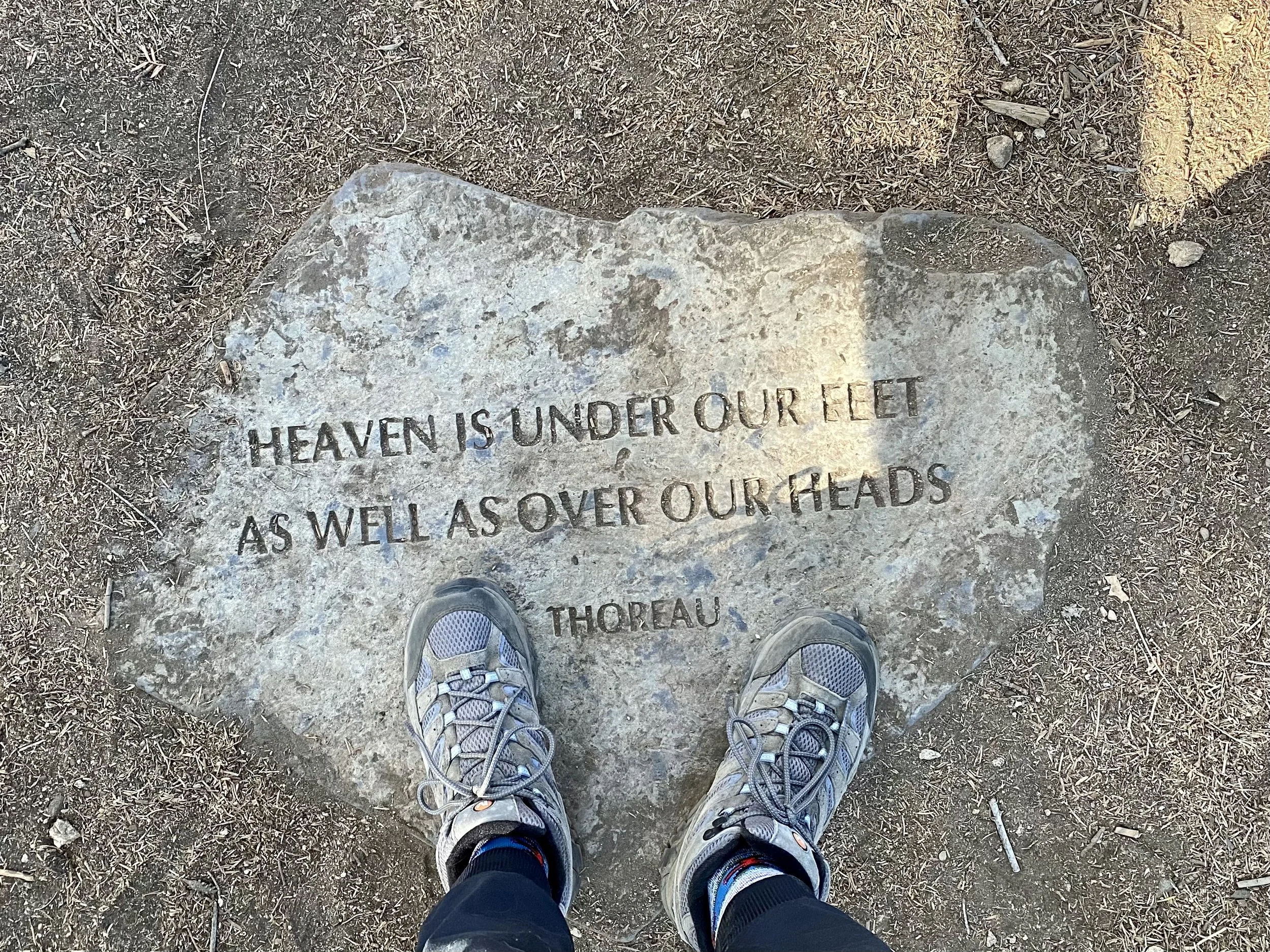 A stone engraved with a quote, seen from above, with a person's feet in running shoes standing on it. The quote reads "HEAVEN IS UNDER OUR FEET AS WELL AS OVER OUR HEADS" by THOREAU.