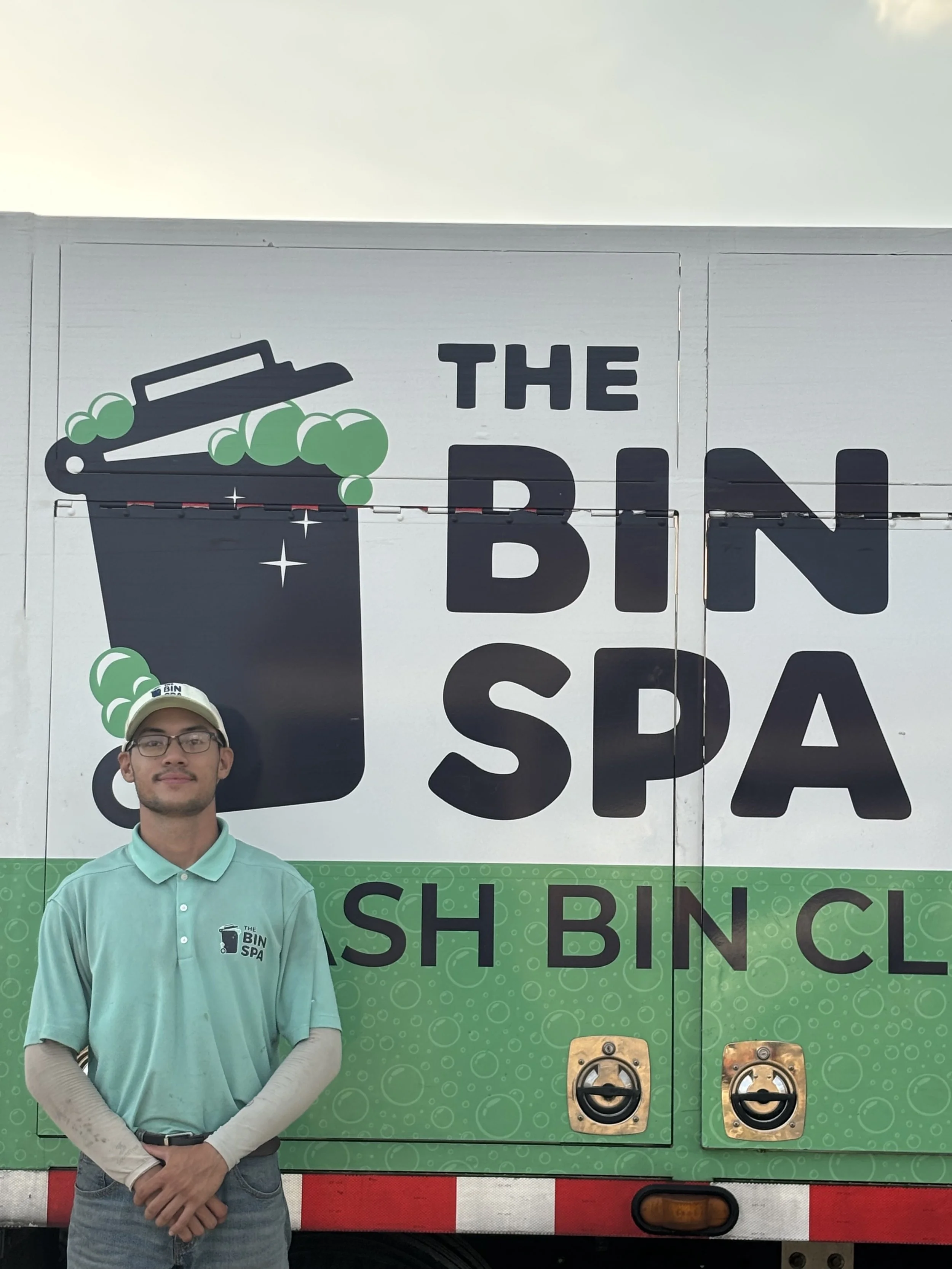 A man wearing glasses, a light green polo shirt, and beige sleeves stands in front of a green and white vending truck for The Bin Spa, a shoe and bag cleaning service, with his hands clasped.