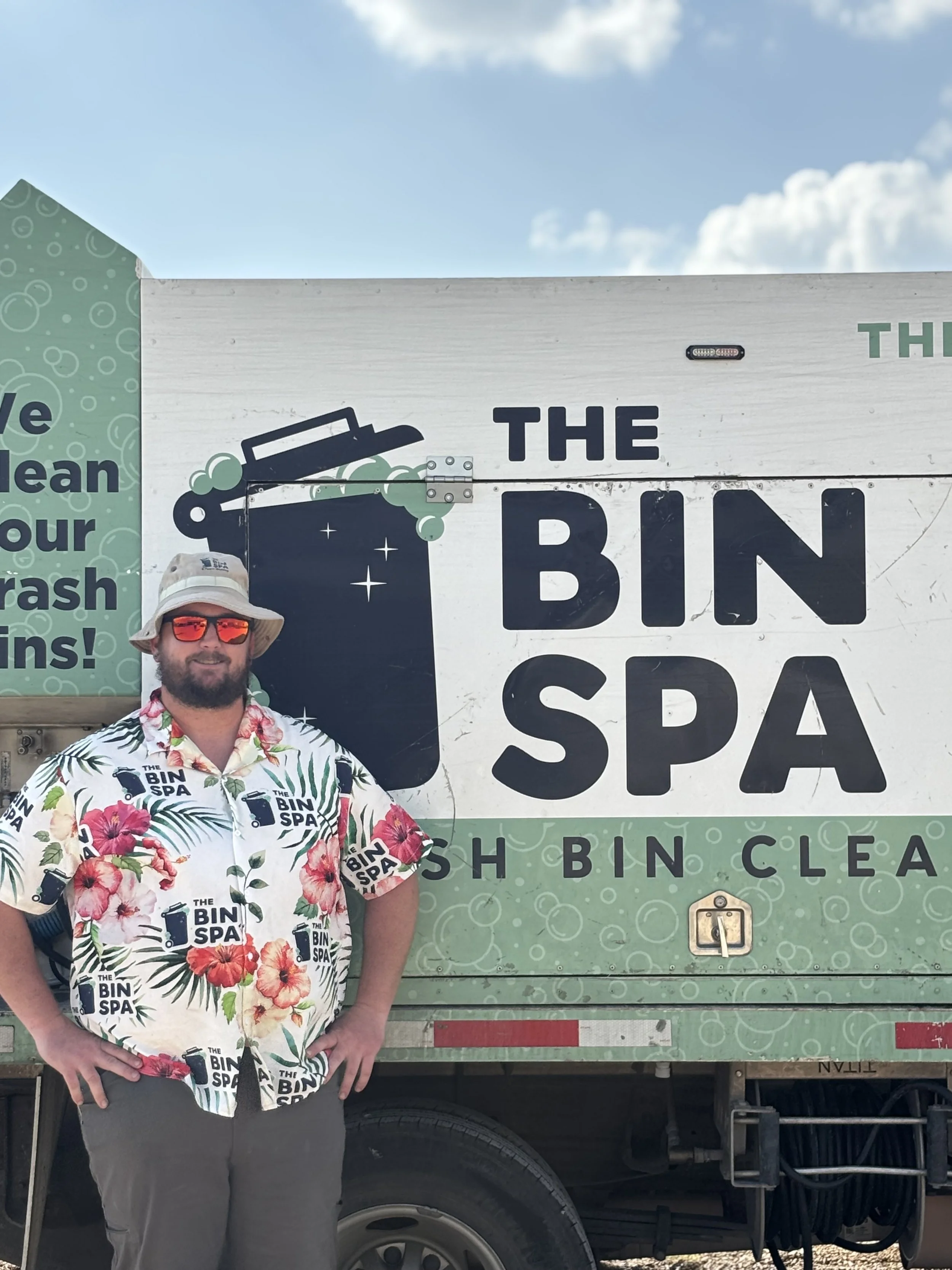 A man wearing a hat, sunglasses, and a shirt with a tropical floral pattern stands in front of a recycling truck. The truck has signage that reads 'The Bin Spa' and promotes trash cleaning.