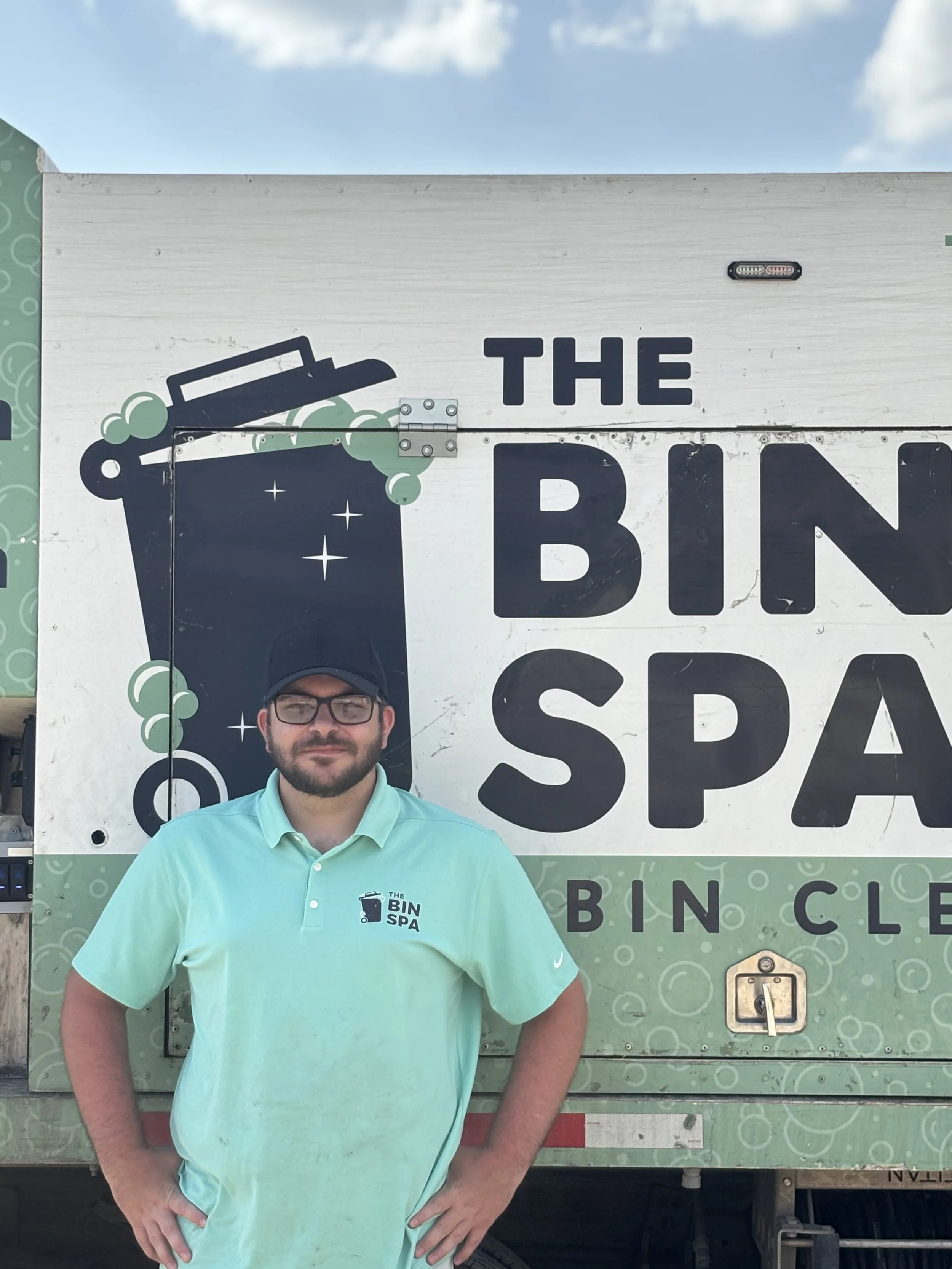 A man in a light blue polo shirt and black glasses standing in front of a green mobile trash receptacle with a logo that says 'The Bin Spa.'