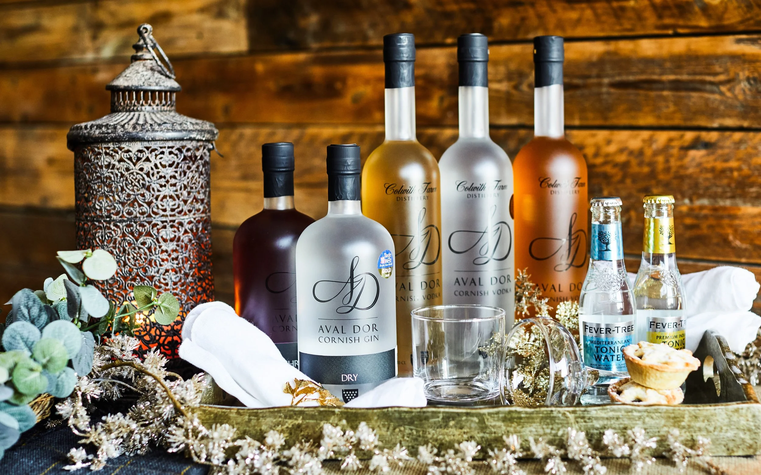 Assorted bottles of Aval Dor Cornish gin and vodka with Fever-Tree tonic water, decorative lantern, greenery, clear glass, and pastries on a wooden surface.