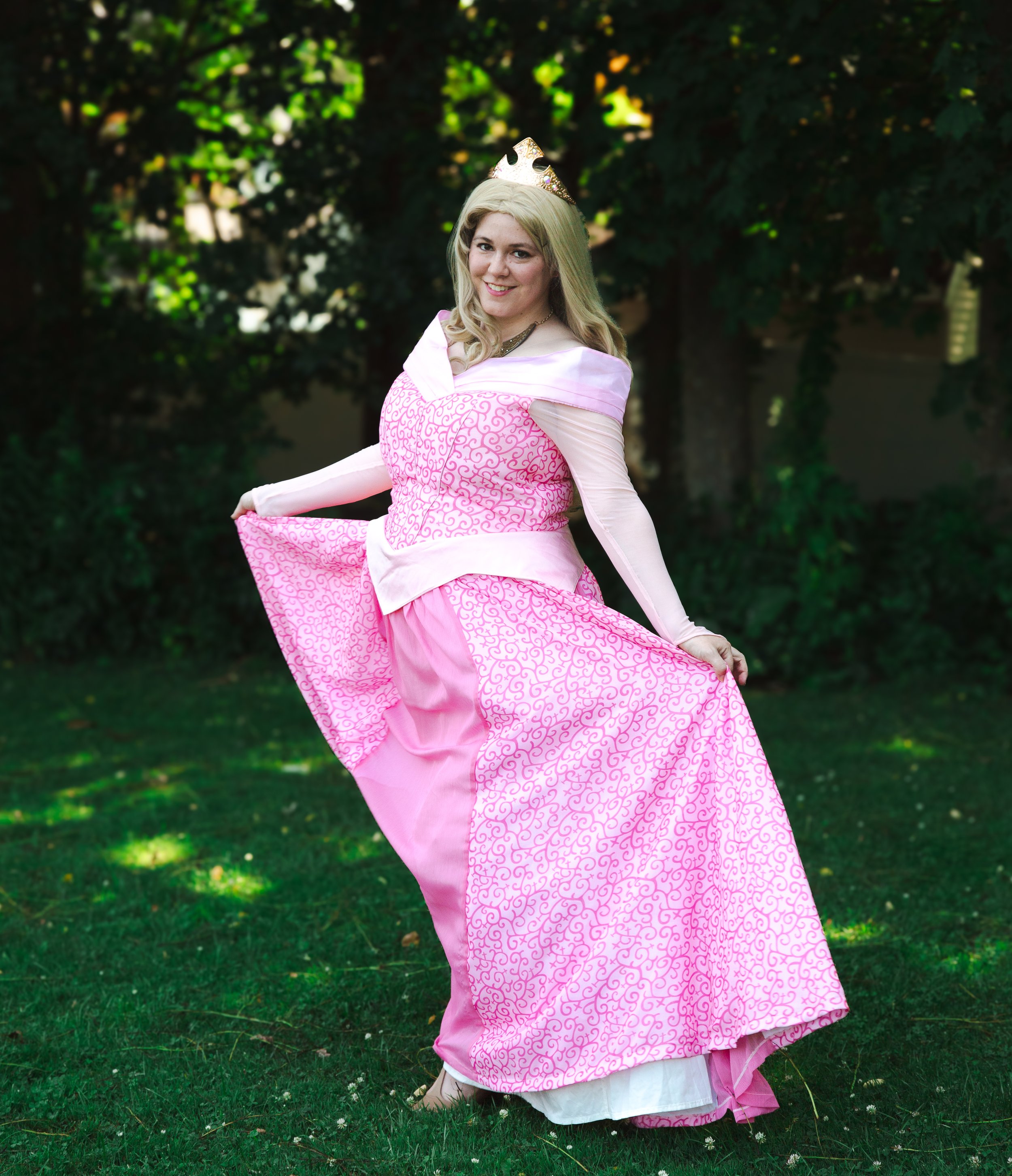 Sleeping Beauty party princess in a pink gown with intricate patterns, wearing a golden crown, standing outdoors on grass with green trees in the background, smiling and holding out her dress.