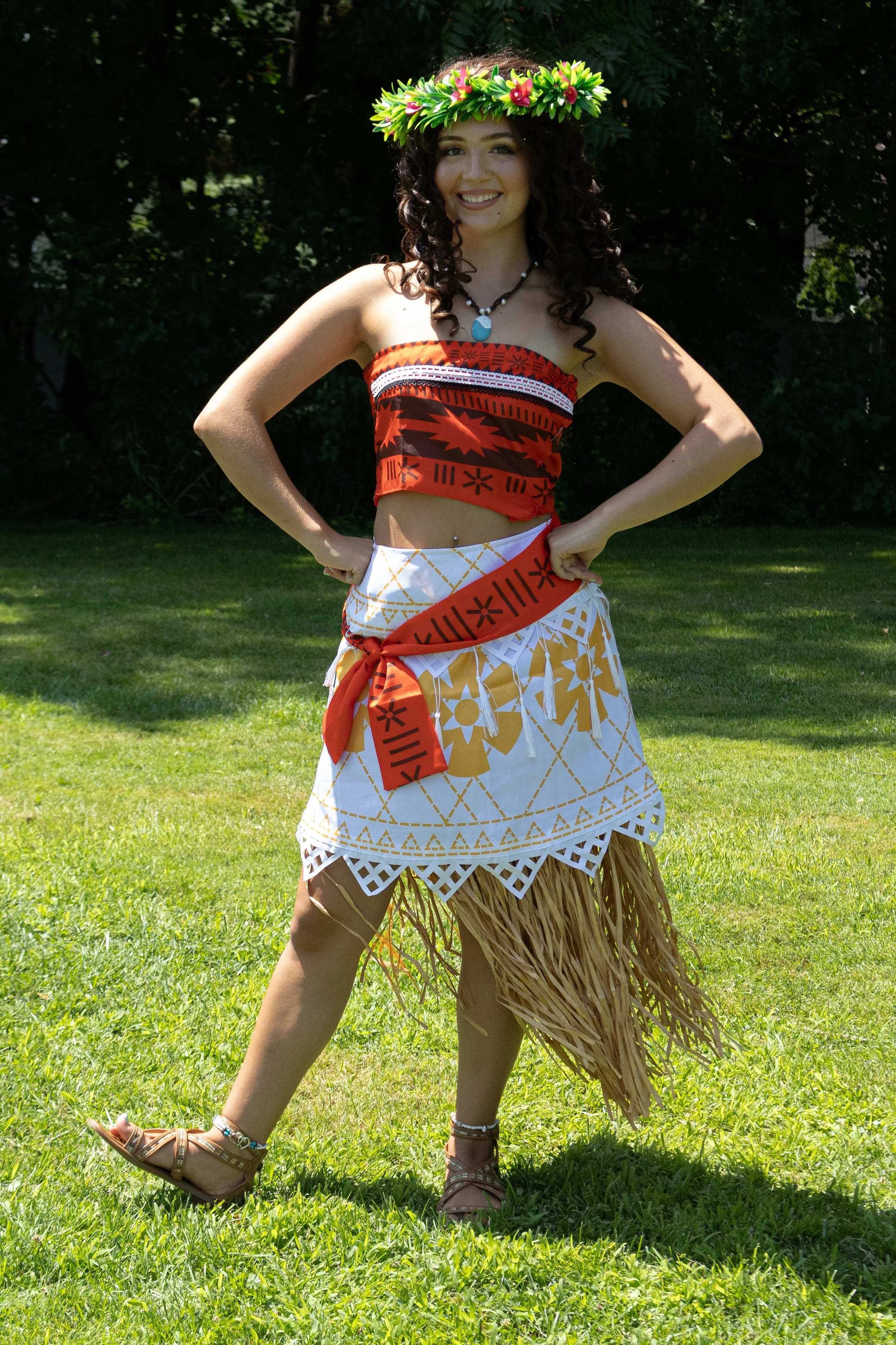 Island Voyager party princess dressed in a red, black, and white traditional Polynesian costume, wearing a leaf crown, standing in a green, wooded outdoor area, with one arm raised and a smile.