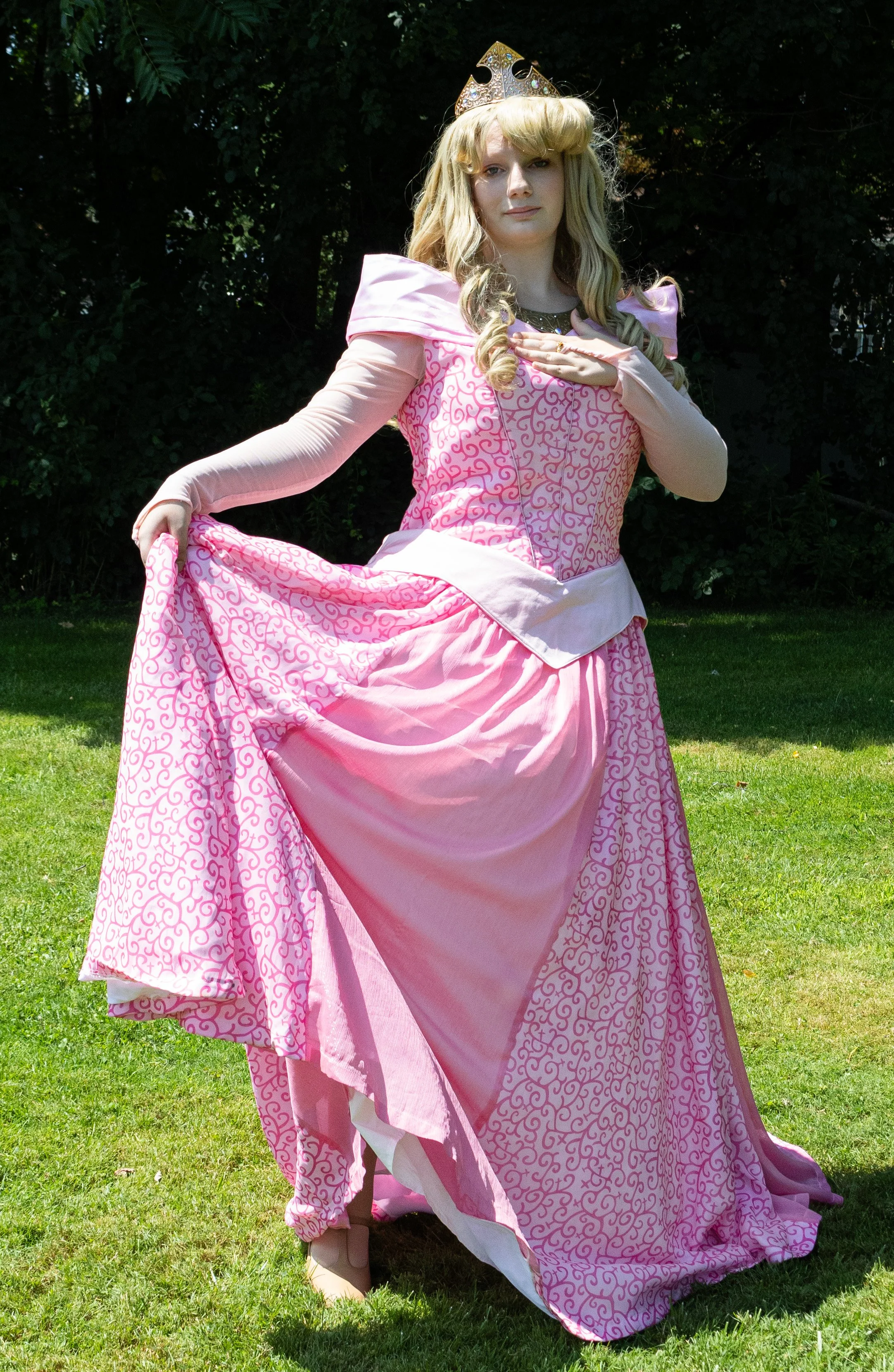 Sleeping Beauty party princess in a pink gown with intricate patterns, wearing a golden crown, standing outdoors on grass with green trees in the background, smiling and holding out her dress.