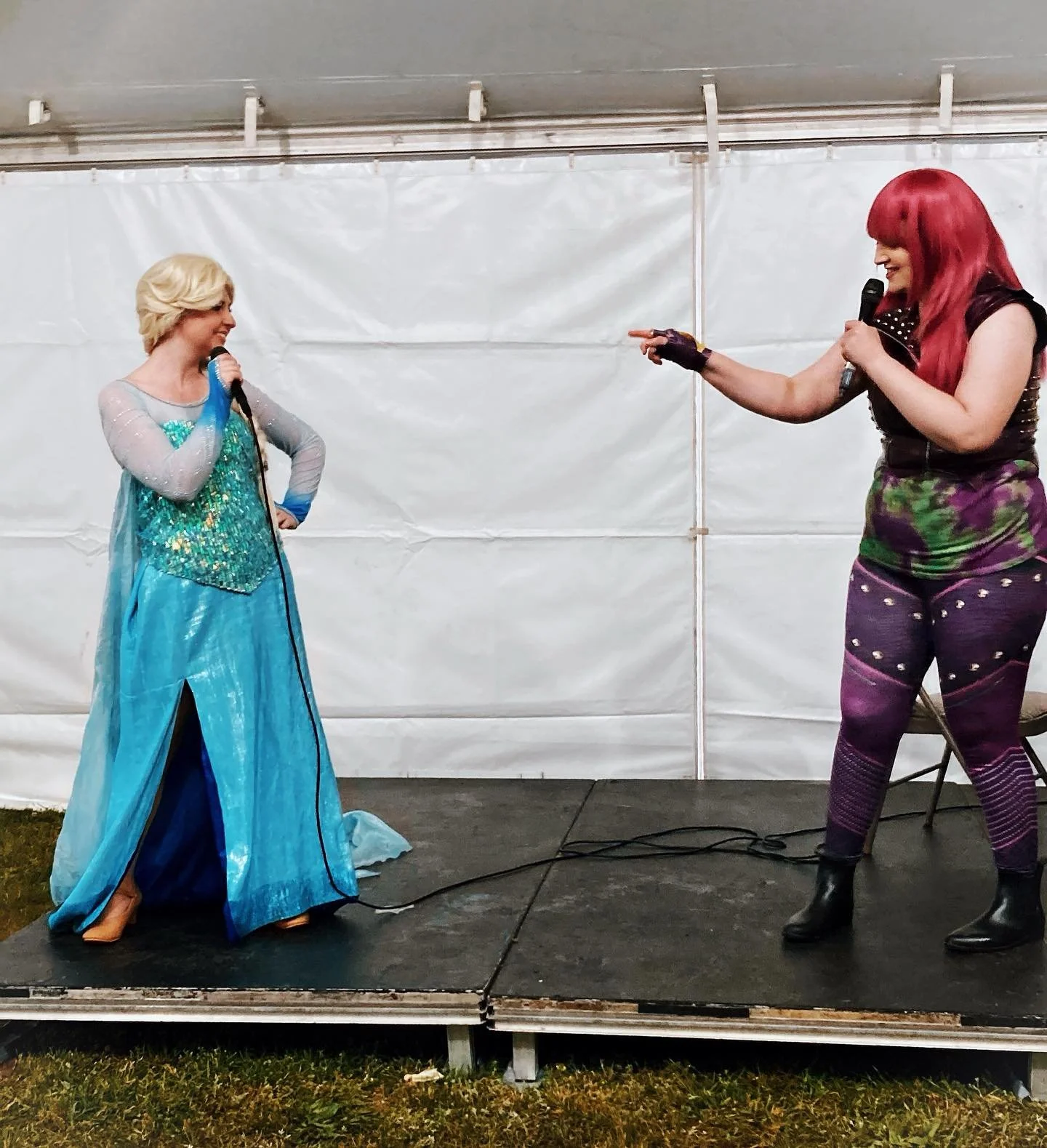 Two Party Princesses perform on a small stage with a white backdrop. The Snow Queen, holding a microphone, and Malicious Teen Villain, also holding a microphone, and pointing at the other woman.