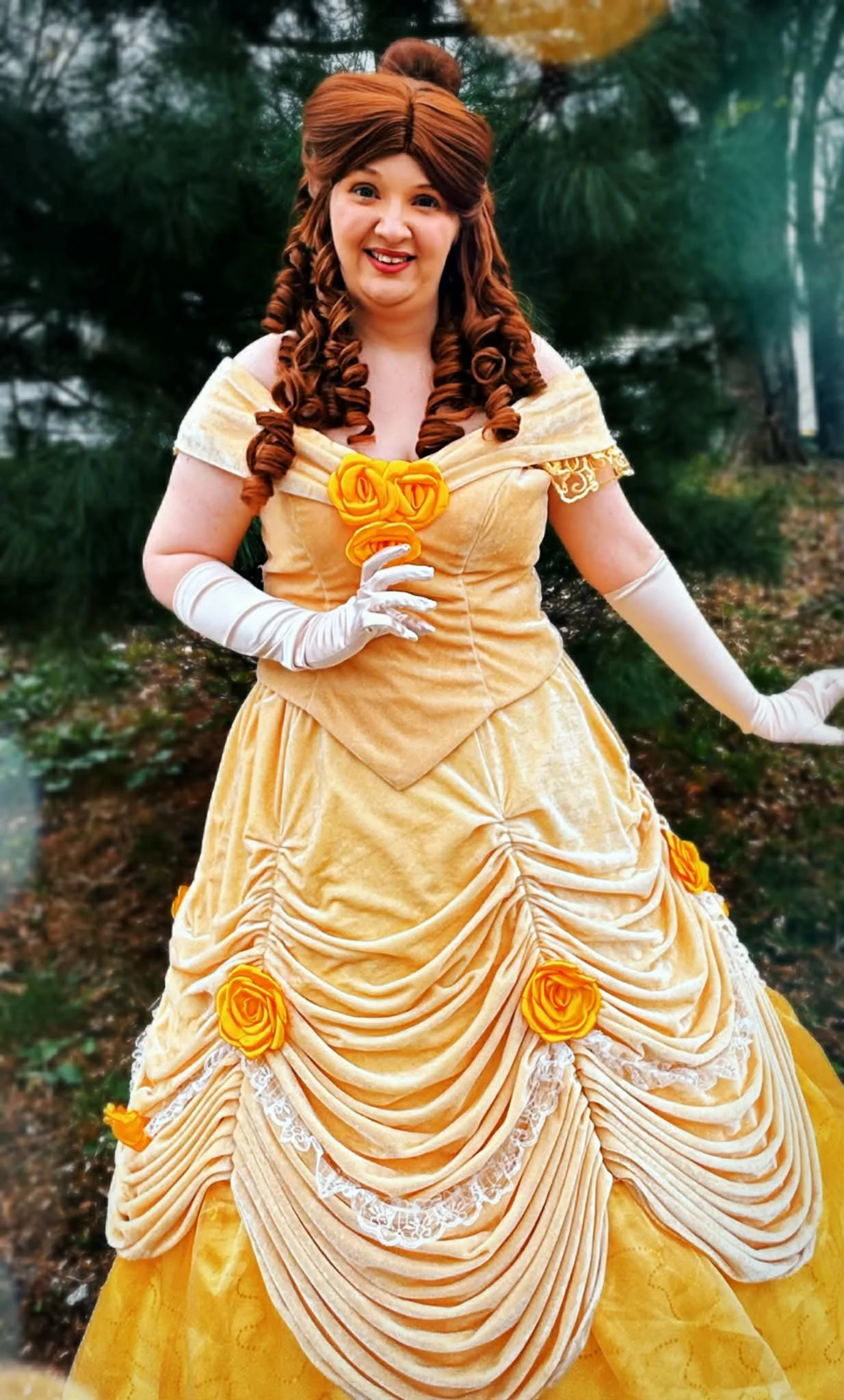 Princess Beauty party princess in a yellow Victorian-style dress with off-shoulder sleeves, long gloves, curly hair, standing on grass in a garden or park.