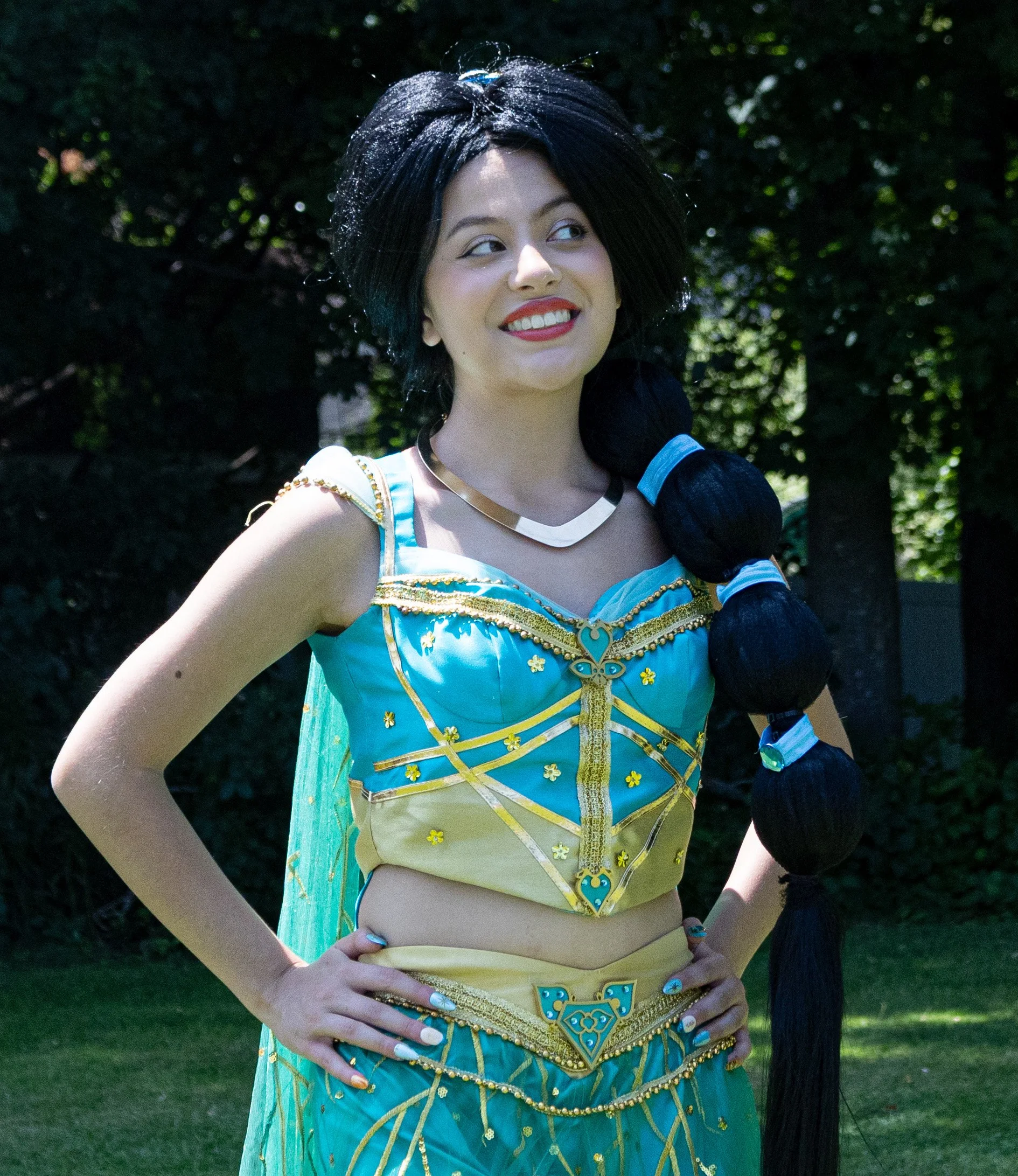 Arabian Princess Party Princess wearing a colorful dress with yellow and green details, stands outdoors in front of green foliage, smiling and crossing her arms.