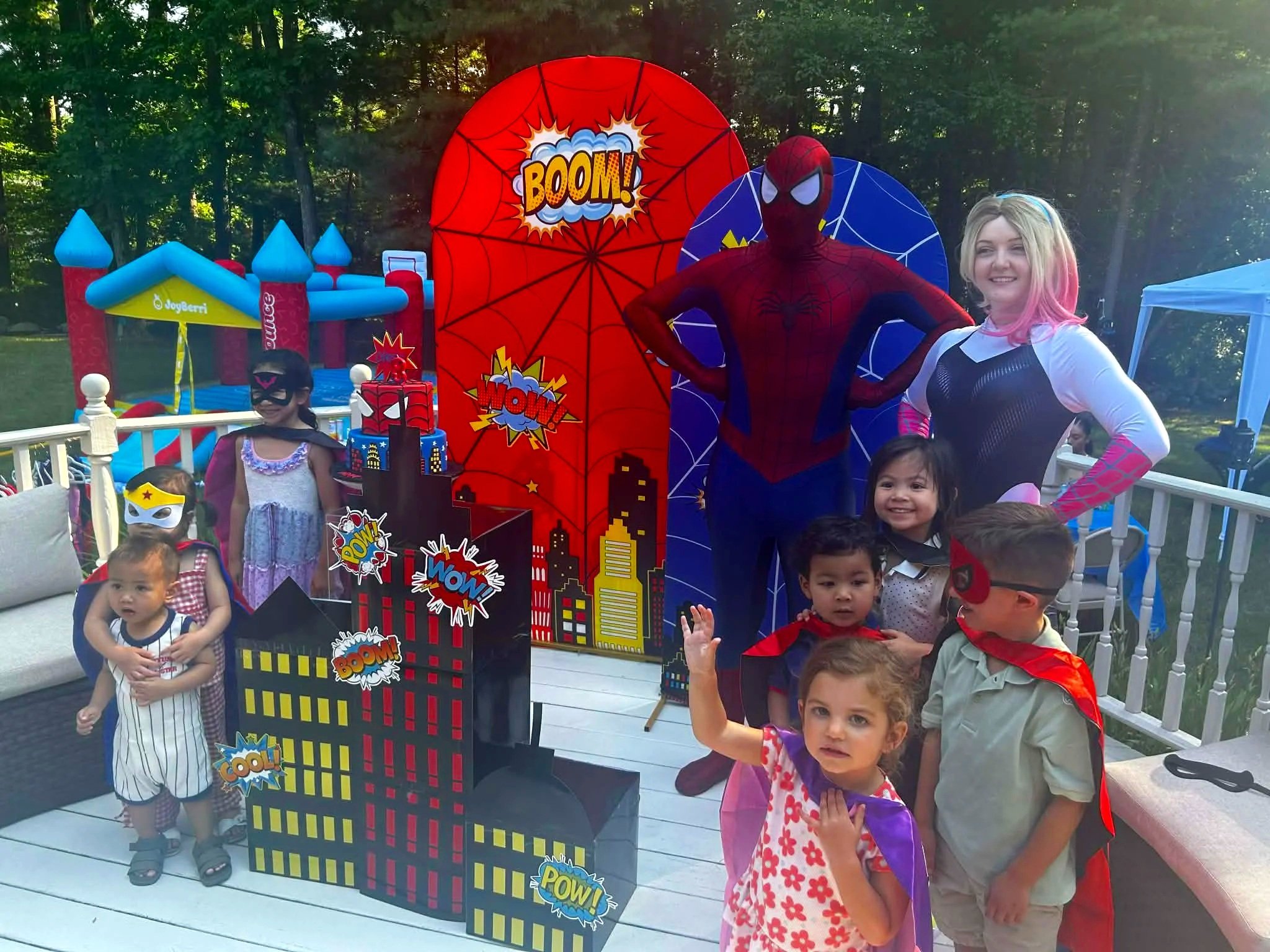 A group of children and Spider-Hero and Ghost Spider-Hero party characters gather outdoors during daytime at a superhero-themed birthday party with colorful decorations, a bounce house, and a cityscape backdrop.
