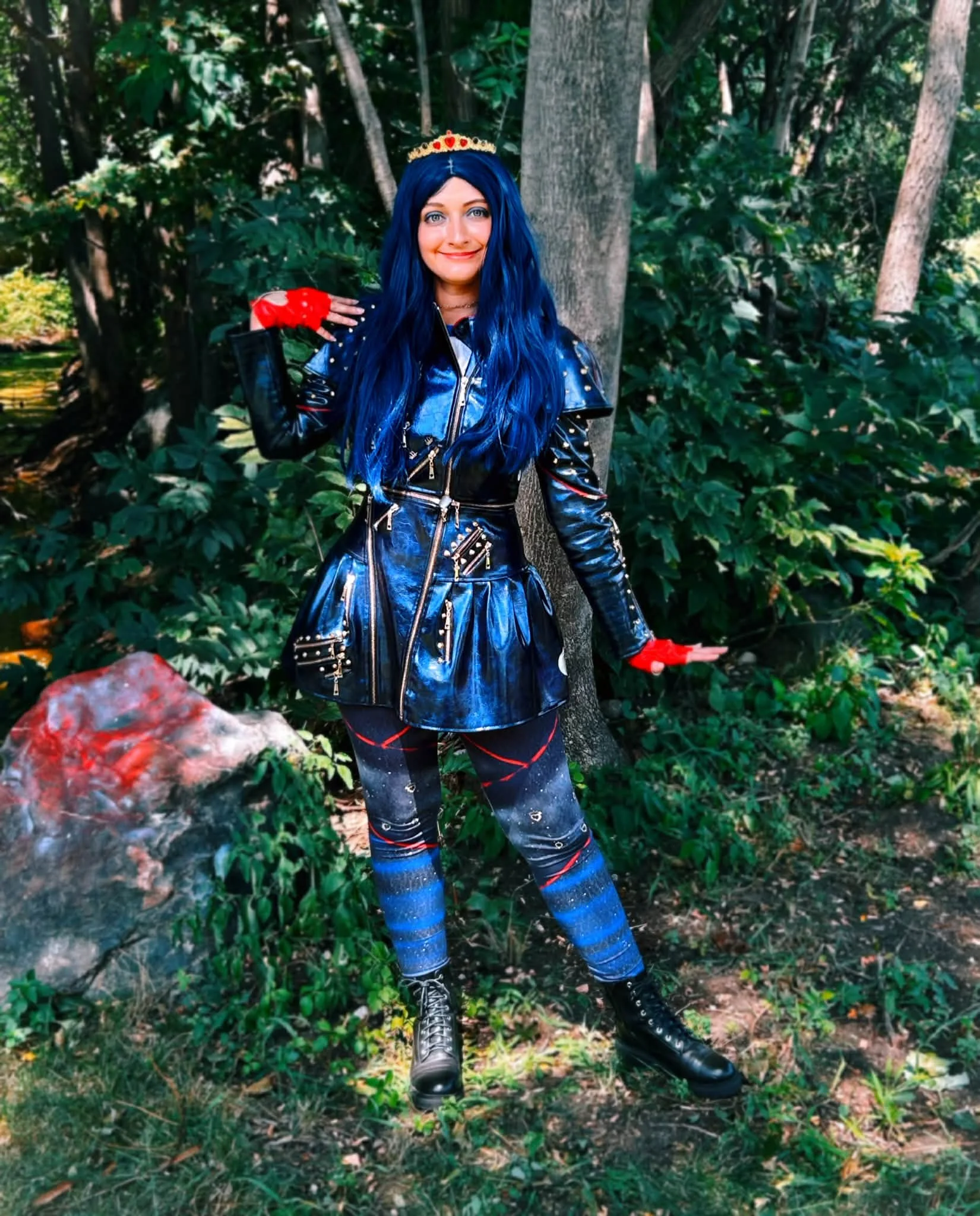 Fashion Queen Teen Villain party princess with long dark blue hair wearing a shiny black leather dress with gold studs, red gloves, and red lipstick, standing against a gray brick wall.