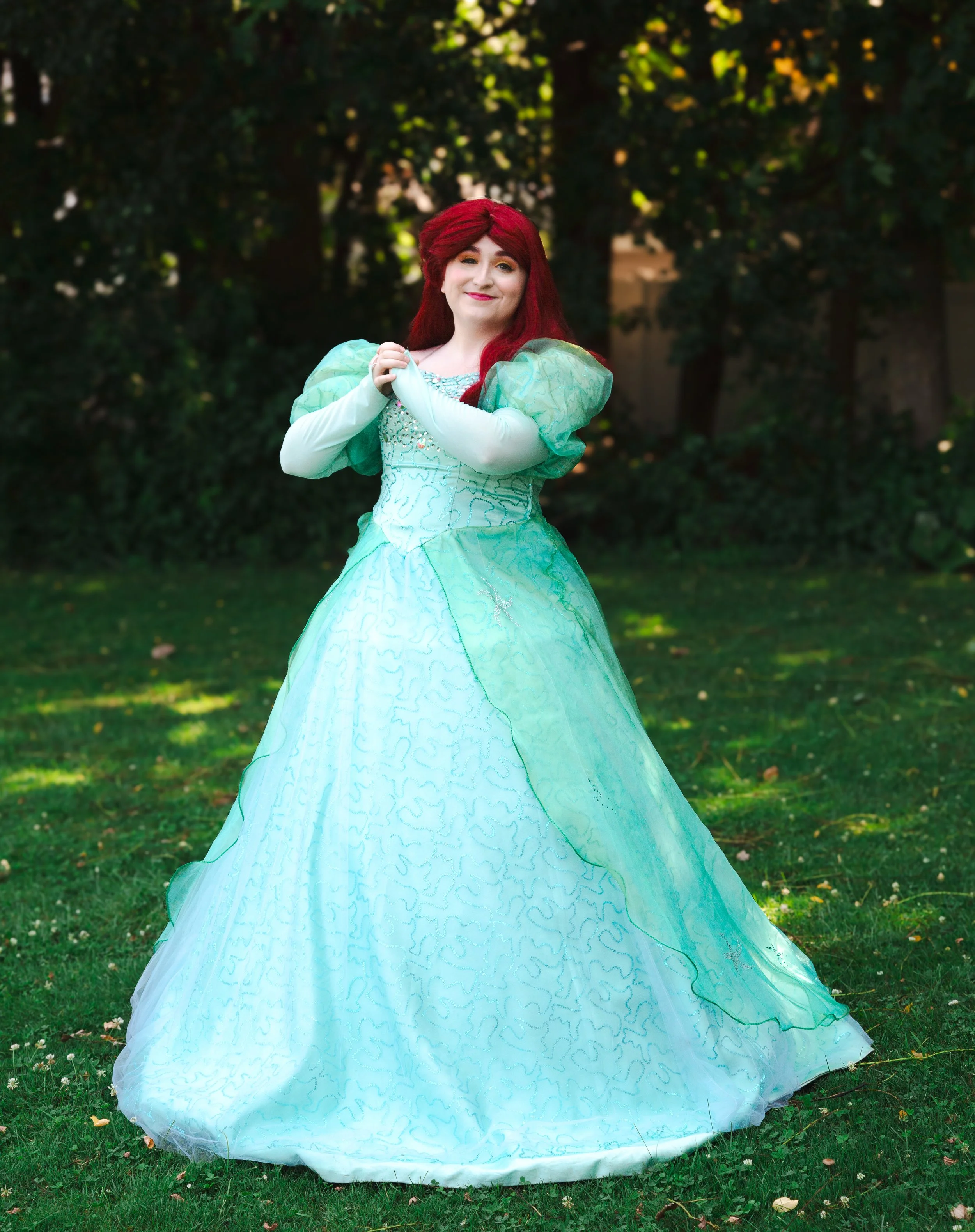 The Little Mermaid party princess standing on grass in front of trees, wearing a light blue gown with green puffed sleeves and long red hair.