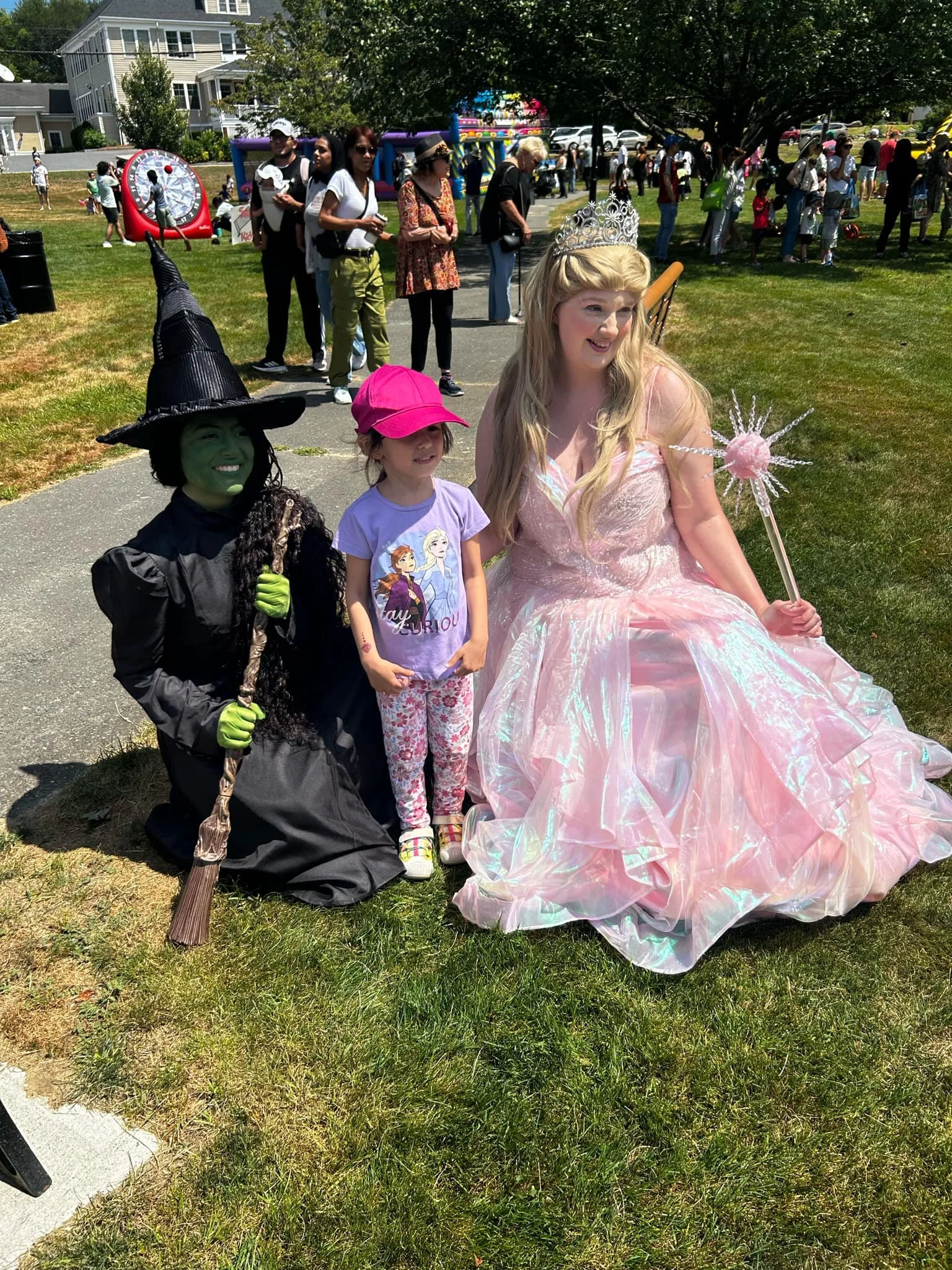 Bubble Witch Party Princess in a pink, iridescent gown with a tiara and wand, sits on the grass with a young girl in casual clothes and the Defying Green Witch Party Princess, at an outdoor event with many people and a bounce house in the background.