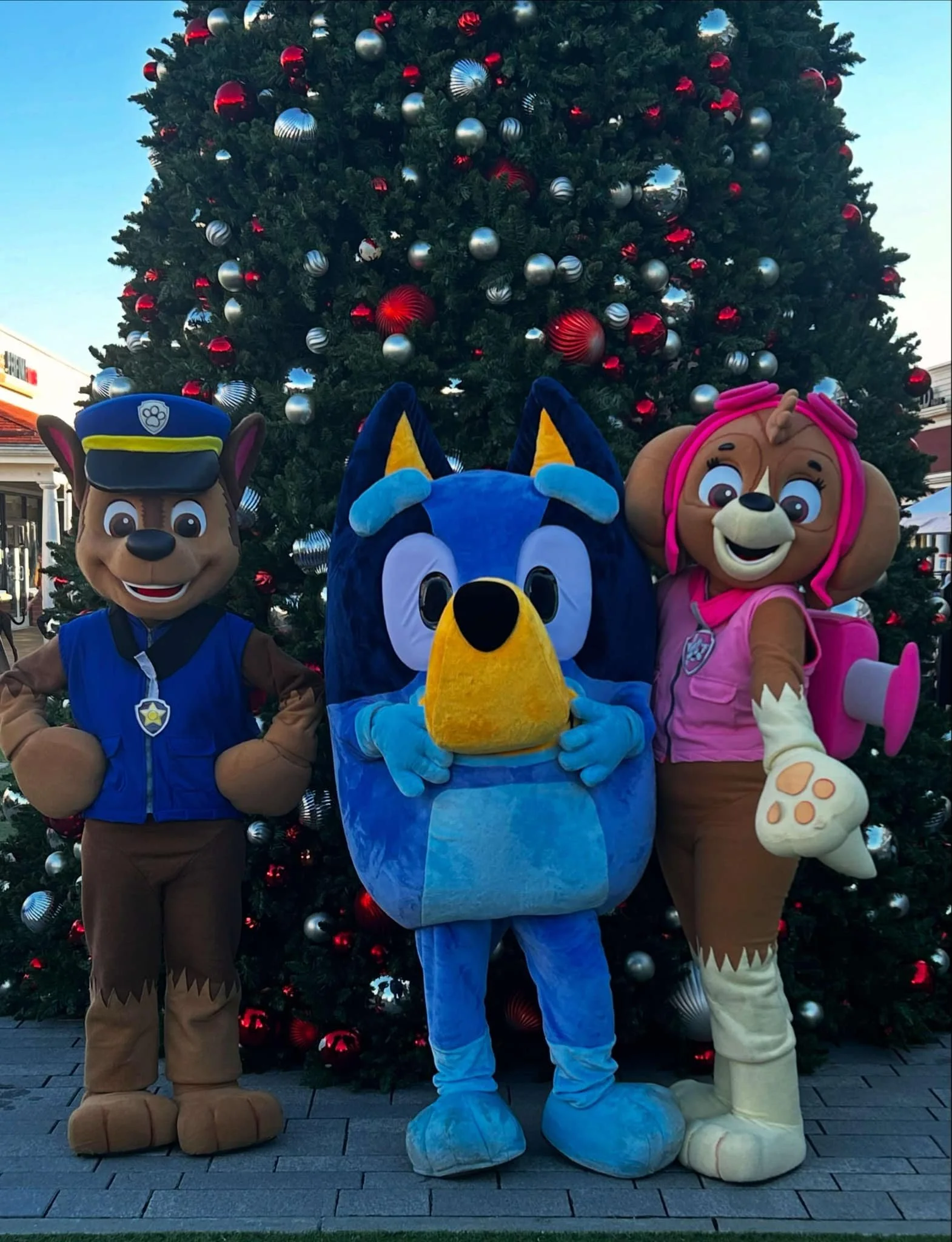 Blue Heeler Puppy, Police Pup, and Aviator Pup party characters posing in front of a decorated Christmas tree with red and silver ornaments.