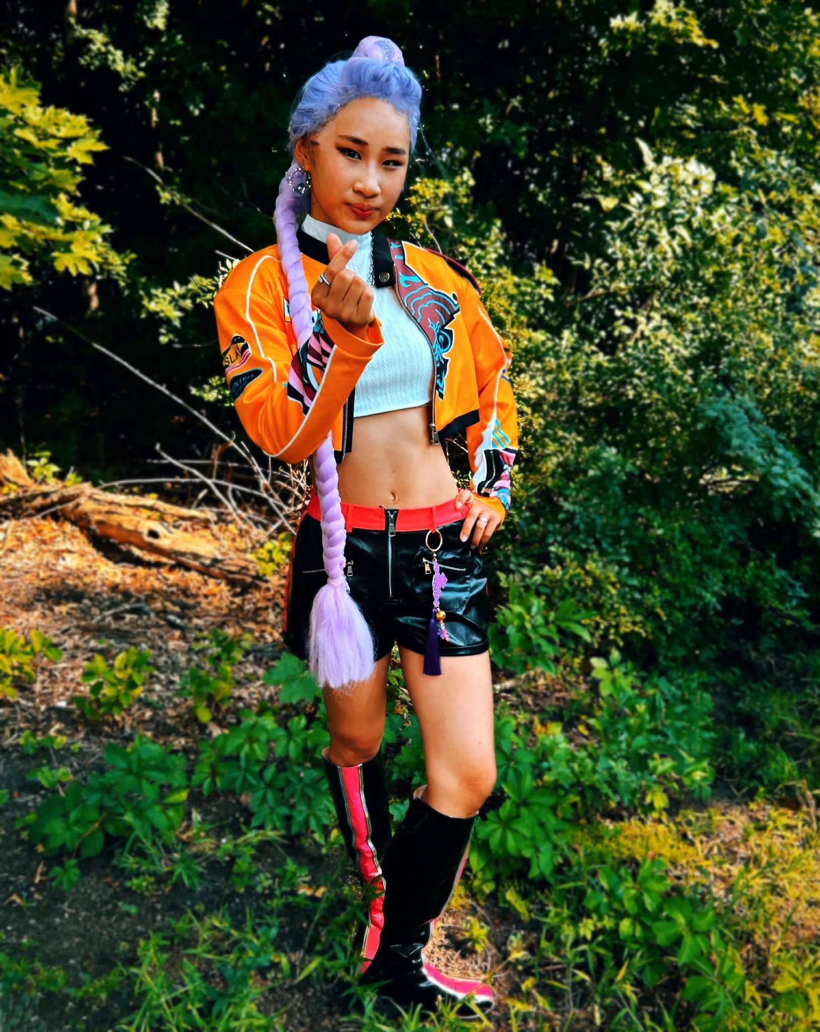 Pop Demon Slayer Party Princess standing outdoors in front of green leafy bushes, dressed in colorful eclectic fashion with vibrant purple hair styled in a long braid, wearing a yellow jacket with patches and black leather shorts with pink trim.
