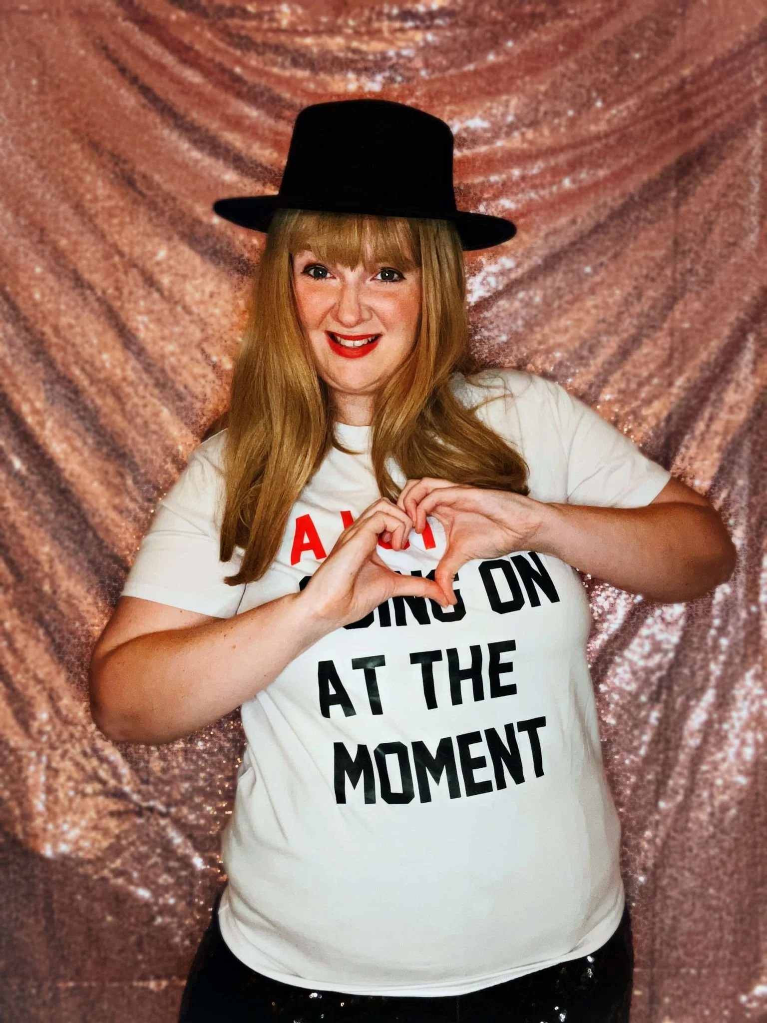 The Eras Queen Party Princess wearing a black hat and white t-shirt with bold black and red text, making a heart shape with her hands against a shimmery pink background.