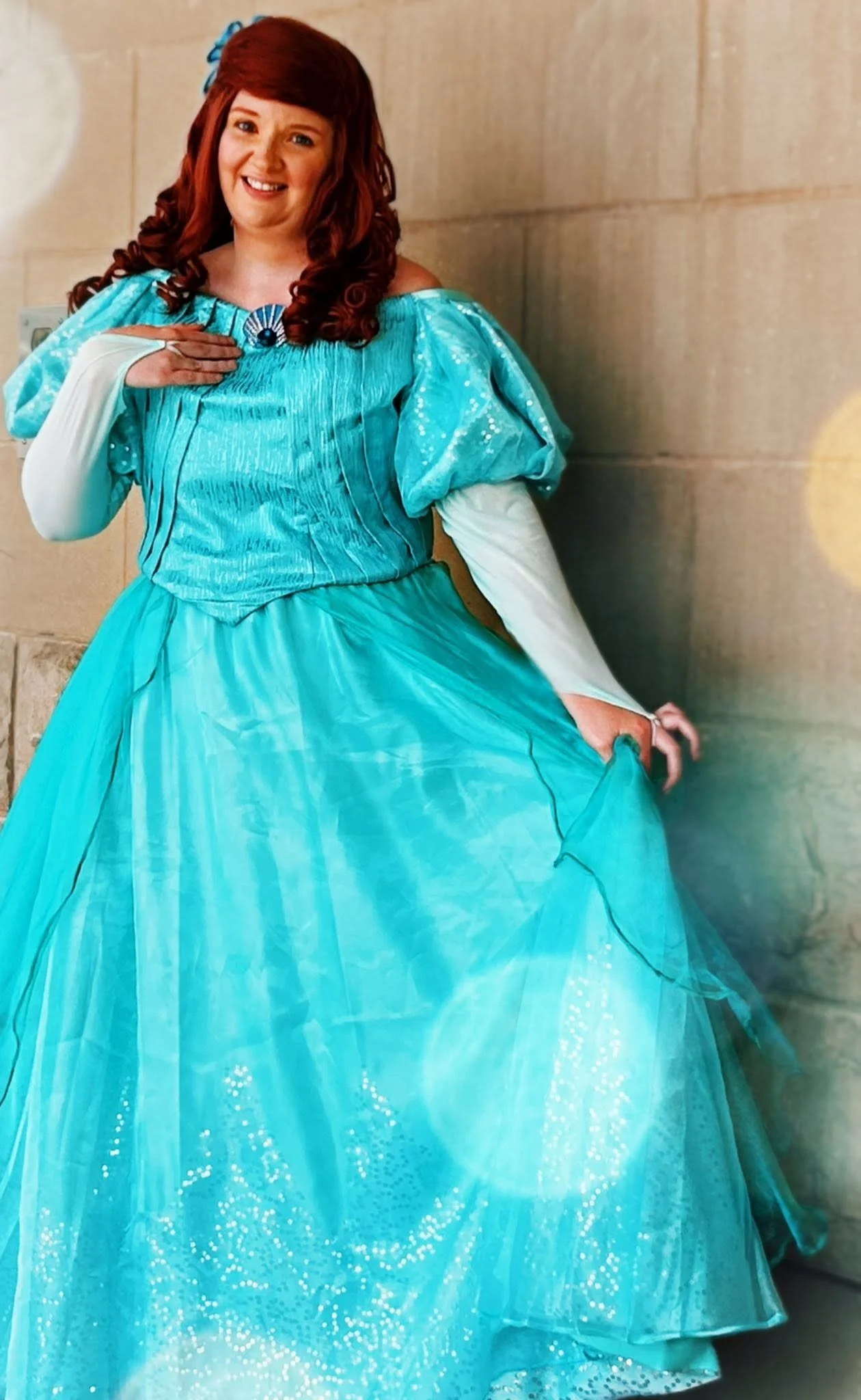 The Little Mermaid party princess standing on grass in front of trees, wearing a light blue gown with green puffed sleeves and long red hair.