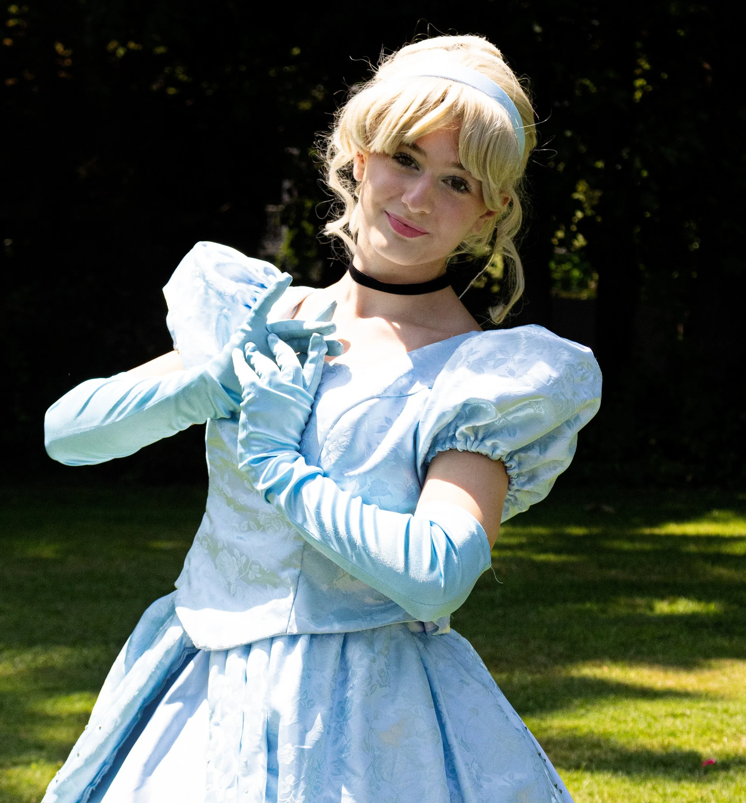 Cinderella party princess dressed in a light blue vintage ball gown with puffed sleeves, gloves, and a choker, standing outdoors on grass with a background of trees and sunlight.