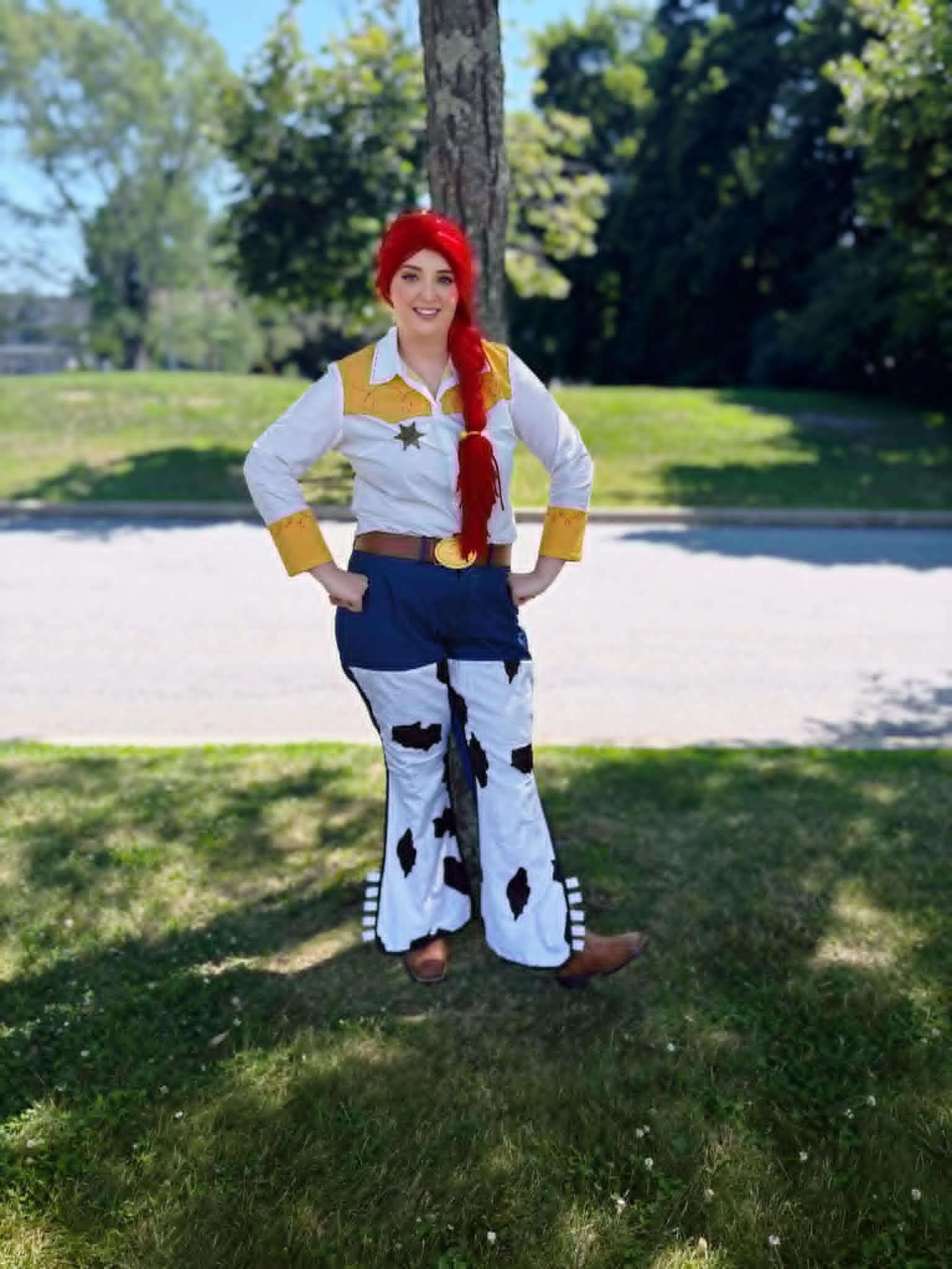 Yodeling Cowgirl Doll party character with red hair standing outdoors on grass in front of a tree, with trees and sky in the background.