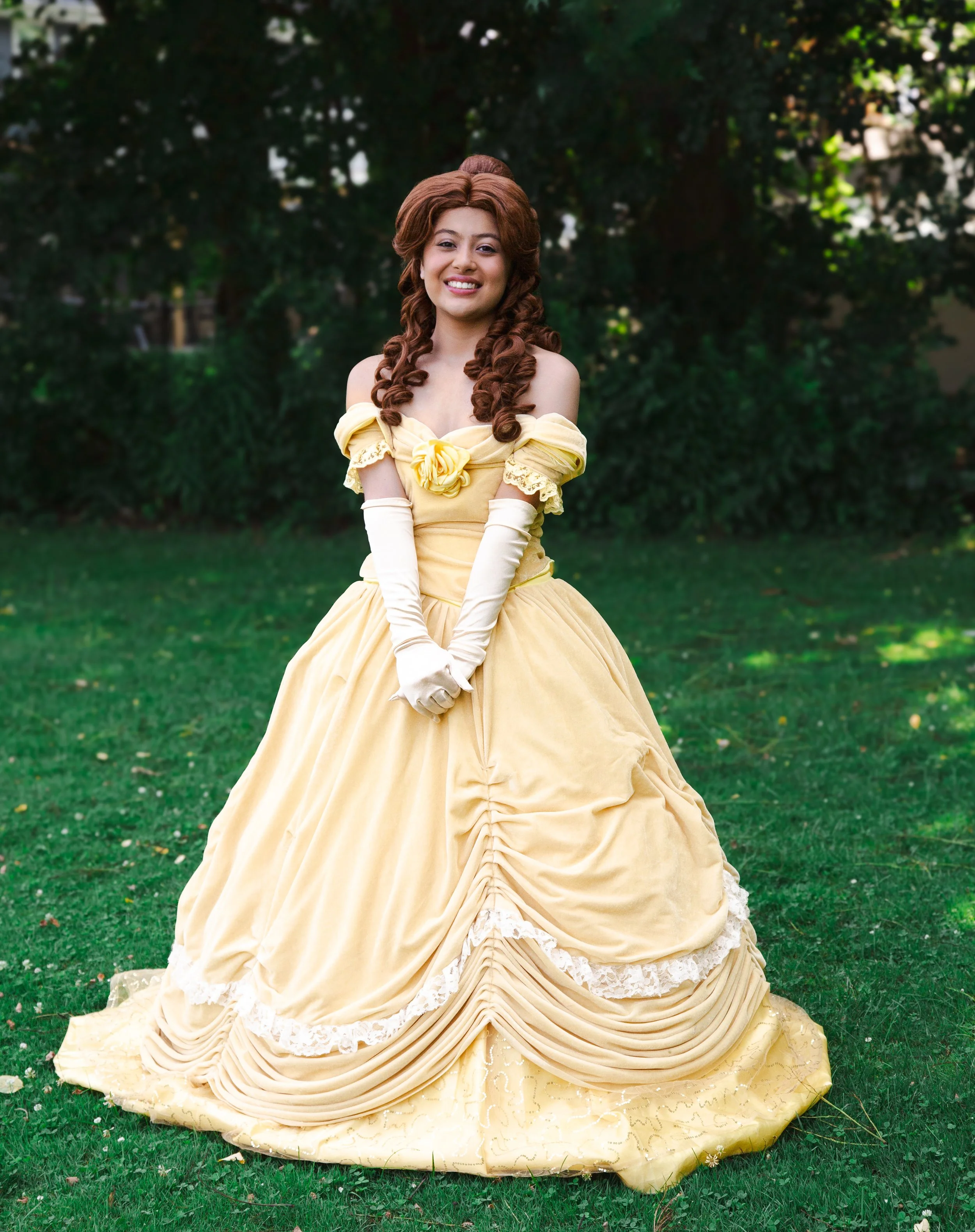 Princess Beauty party princess in a yellow Victorian-style dress with off-shoulder sleeves, long gloves, curly hair, standing on grass in a garden or park.