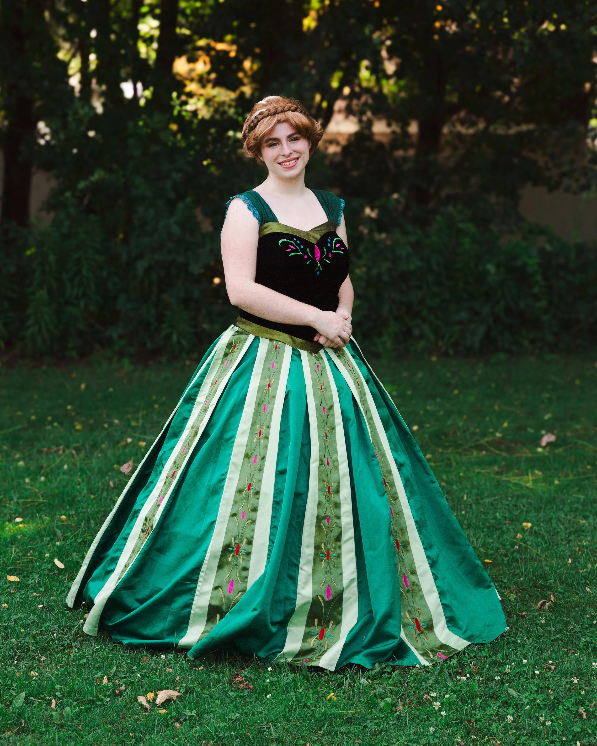Ice Princess in colorful, ornate dress standing on grass with trees in background.
