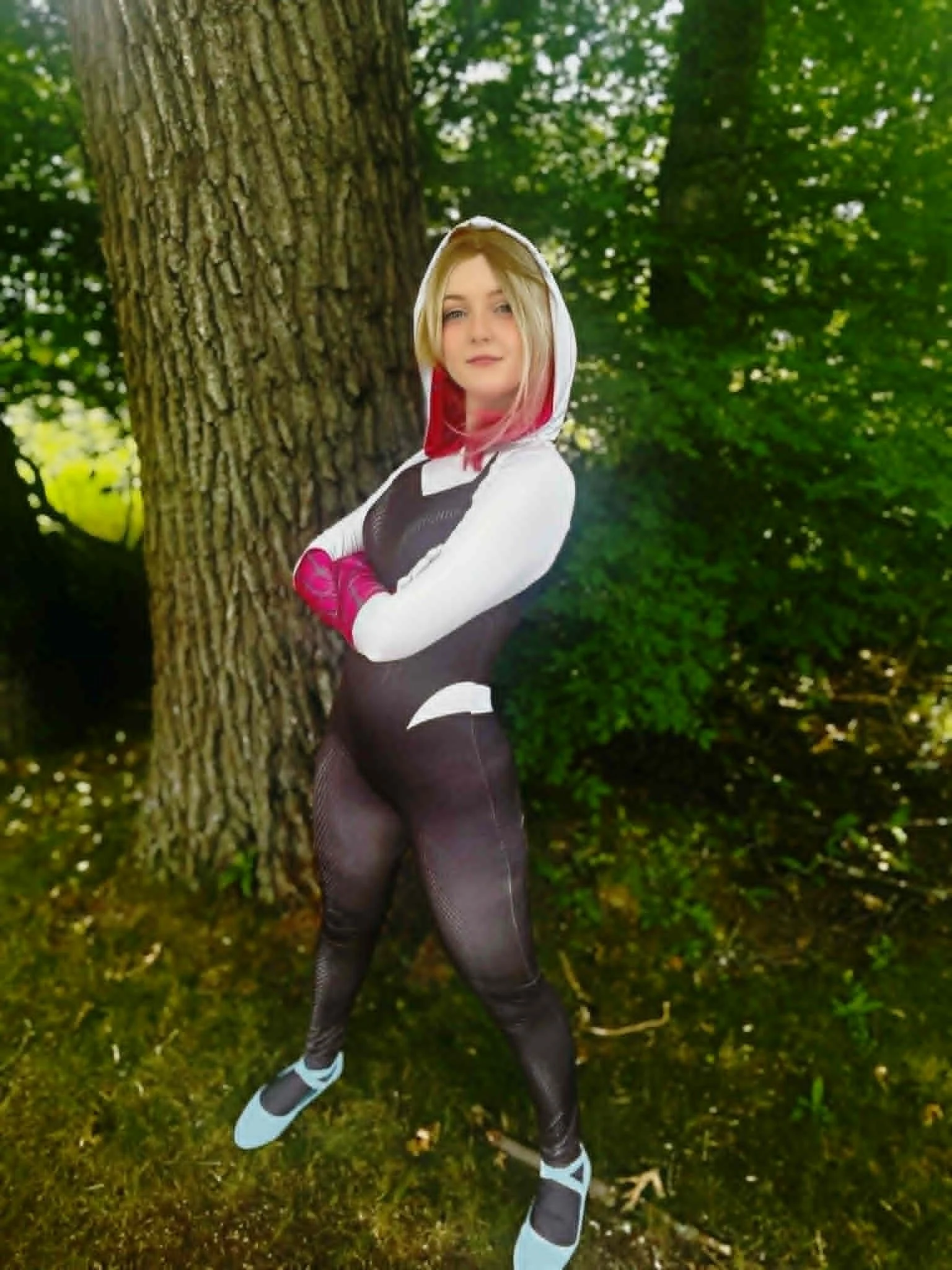 Ghost Spider party character with blonde hair and pink tips wearing a hoodie, black leggings, and gloves leaning against a tree in a forest or park.