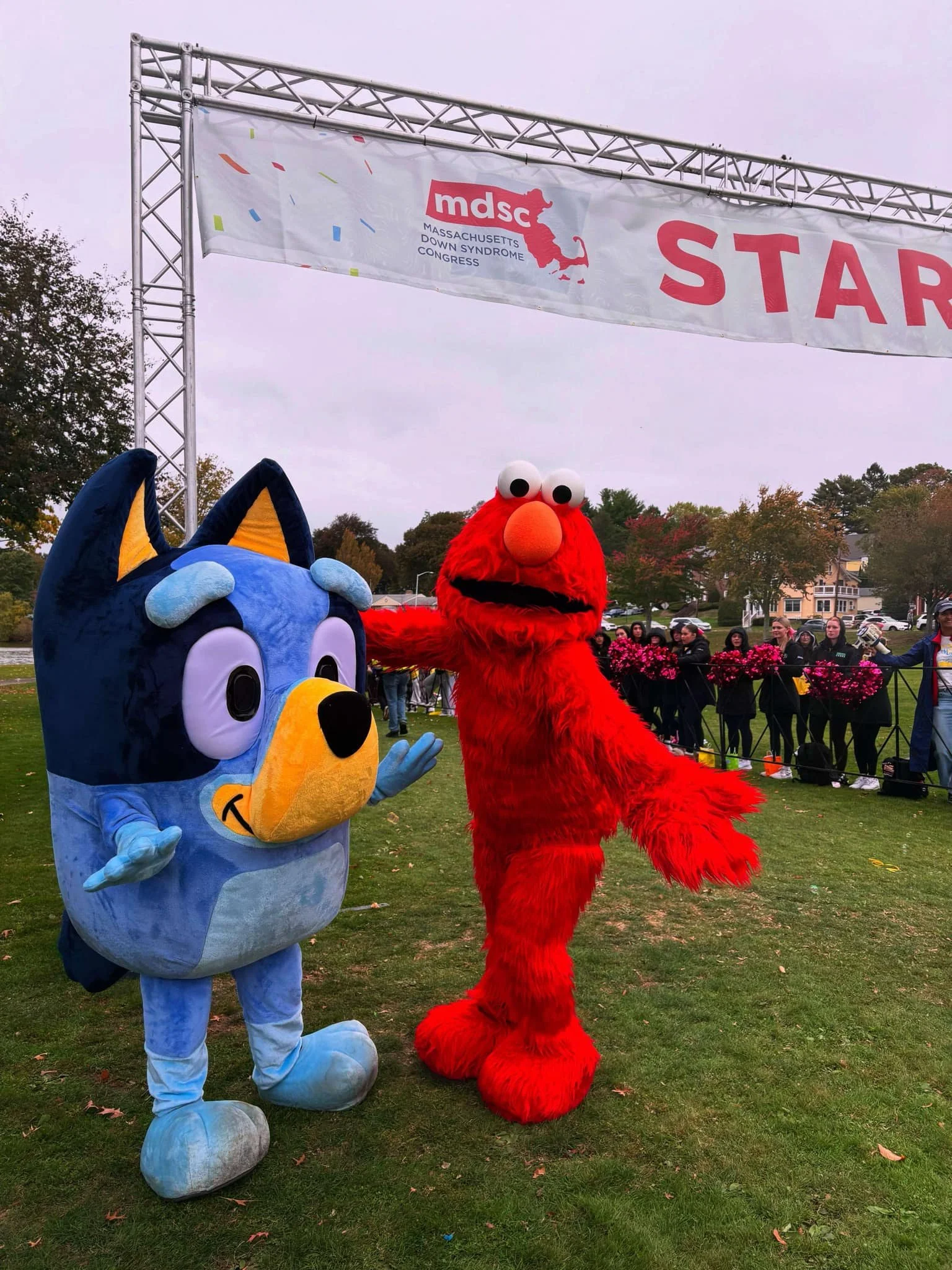 Furry Red Monster and Blue Heeler Puppy party characters posing together at a park event for the Massachusetts Down Syndrome Congress, with a start line banner overhead and people in the background.