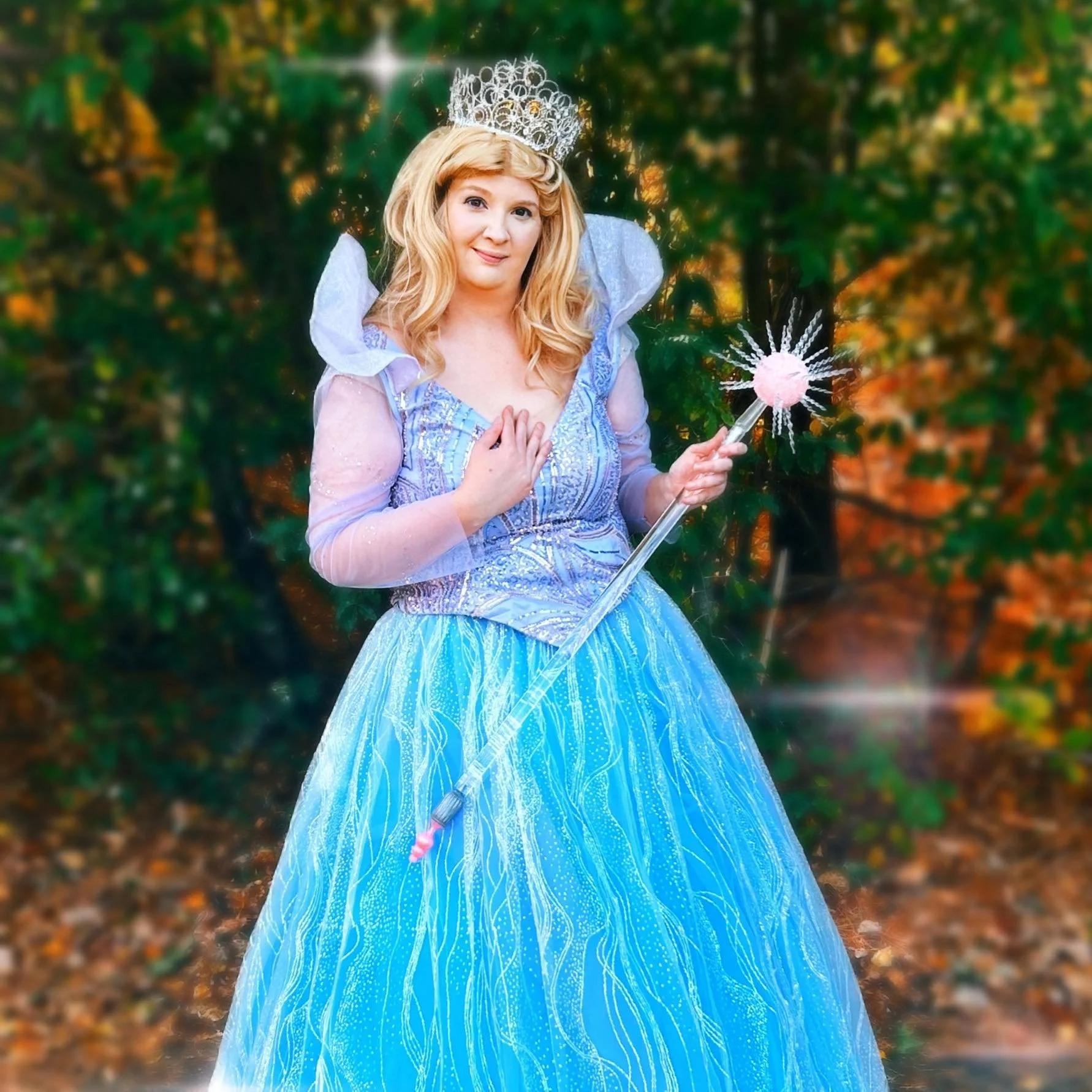 Bubble Witch party princess, wearing a light blue gown with silver detailing and puffed sleeves, standing outdoors in a wooded area with autumn leaves, holding a star-topped wand, and wearing a silver crown.