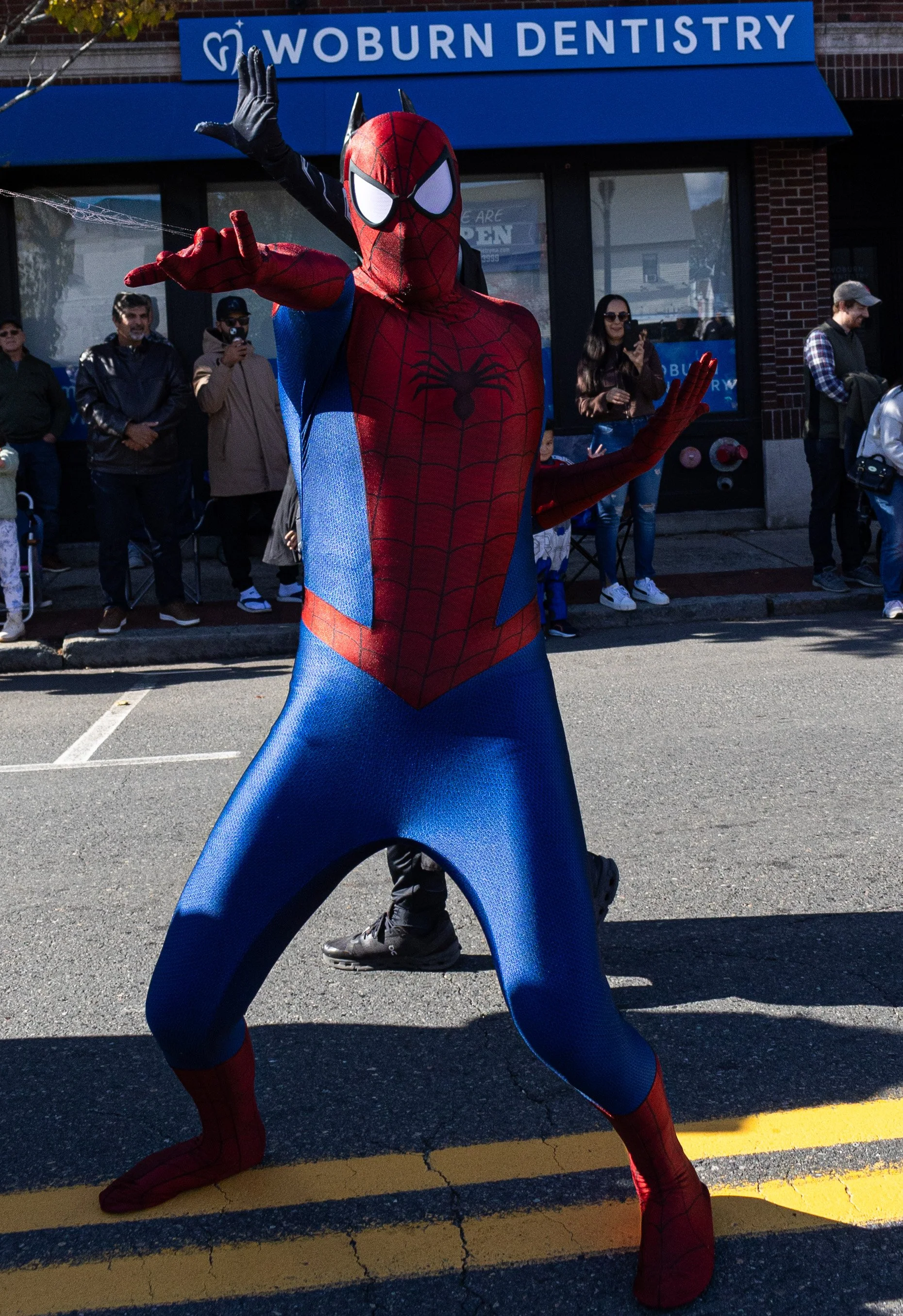 Spider-Hero Party Character posing with a hand gesture on a city street during a public event, with people watching in the background.