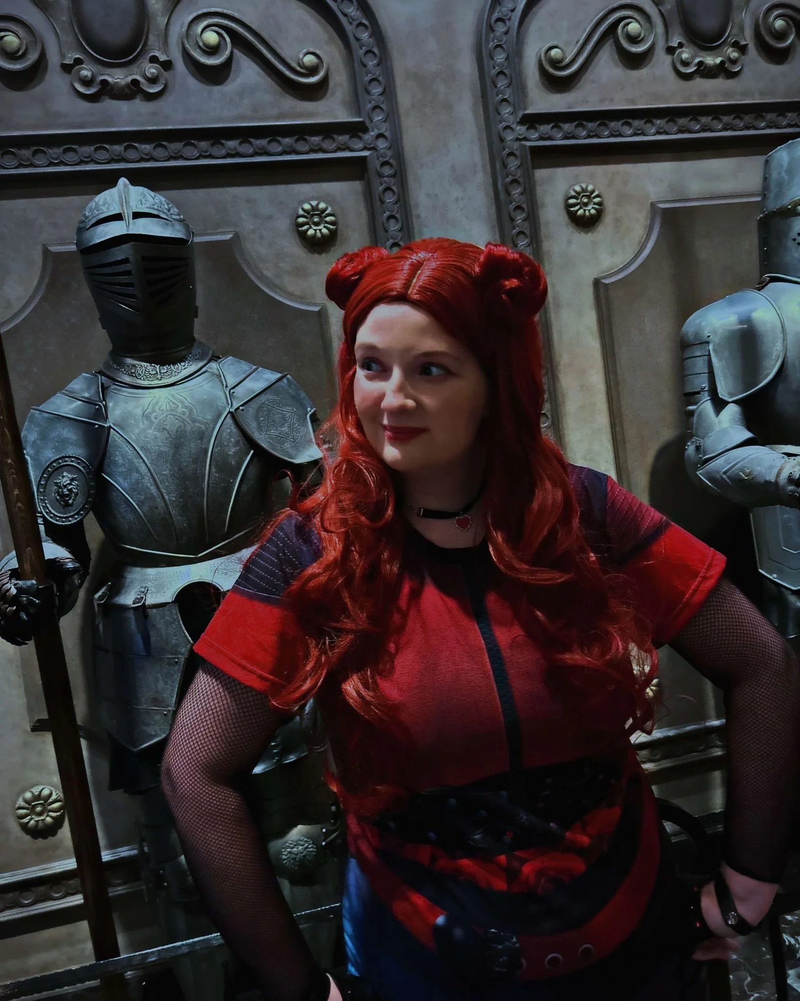 Rebel Teen Villain party character with long red hair styled in buns at the top, wearing a red and black graphic t-shirt, fishnet sleeves, and a choker necklace, standing in front of medieval armor displays.
