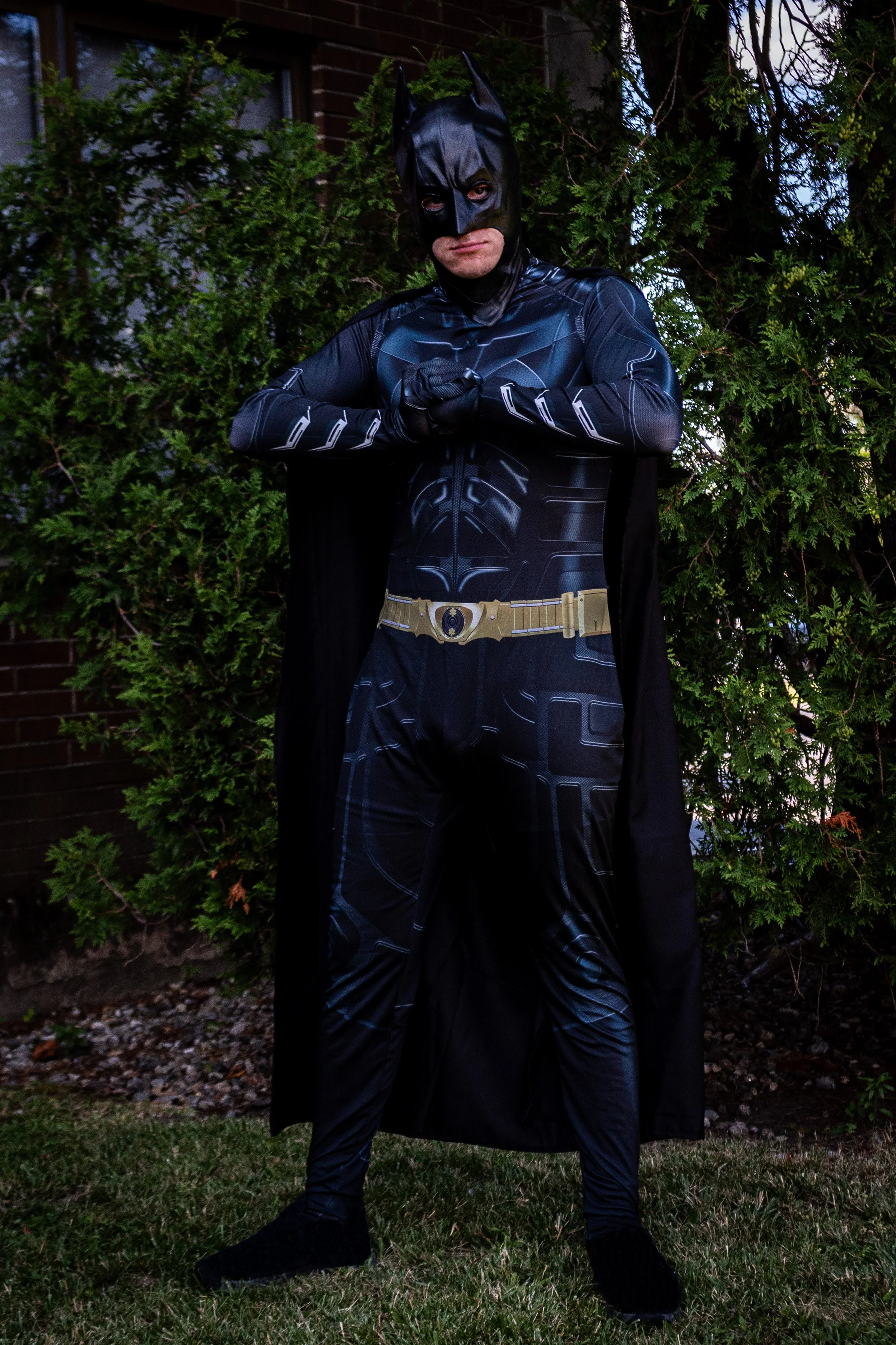 Bat-Hero party character in a suit complete with mask, cape, and utility belt, standing outdoors in front of green bushes.