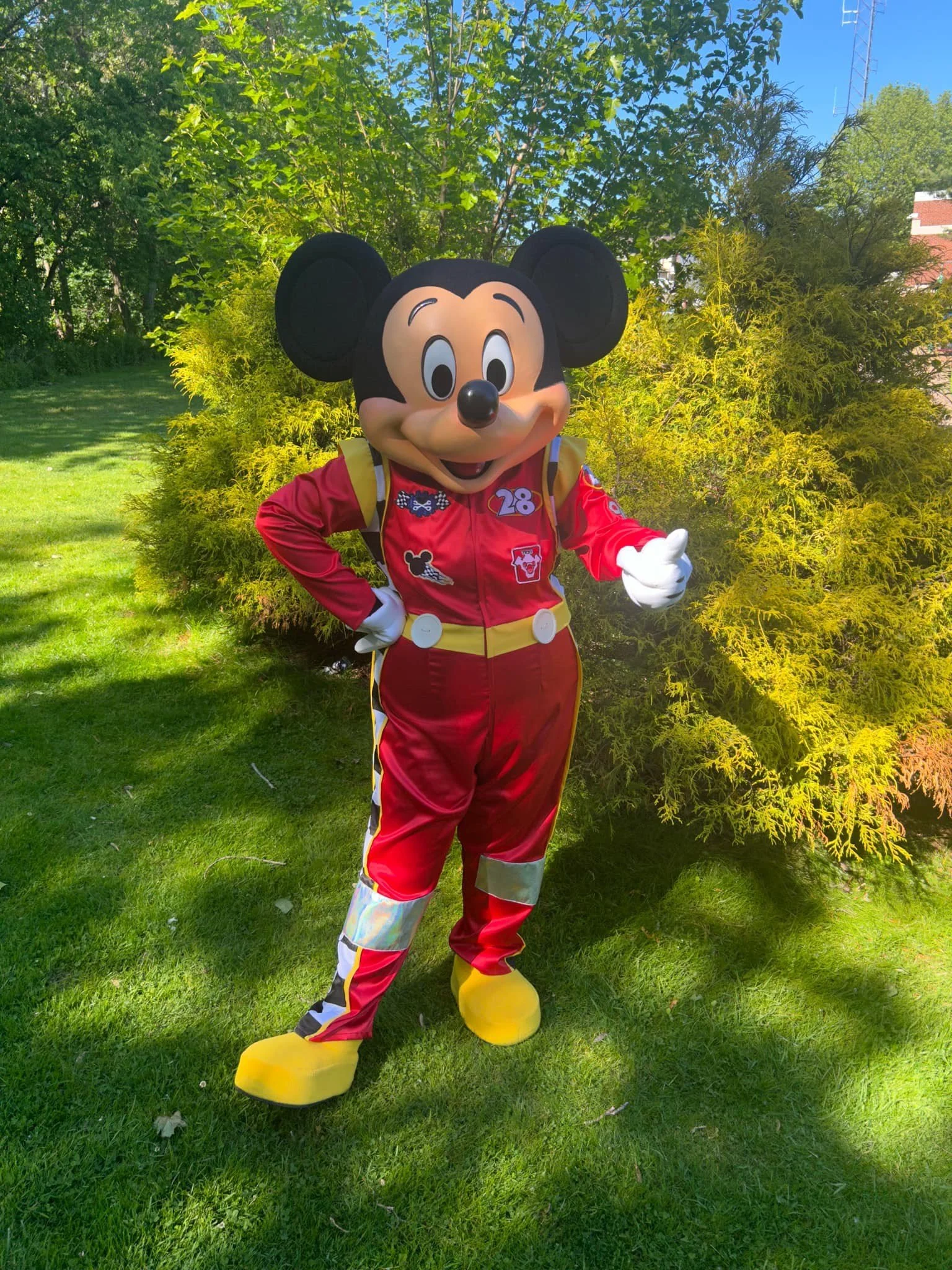 Mr. Mouse party character dressed in a red racing suit with the number 28, standing outdoors on a grassy area with trees and shrubs behind him.
