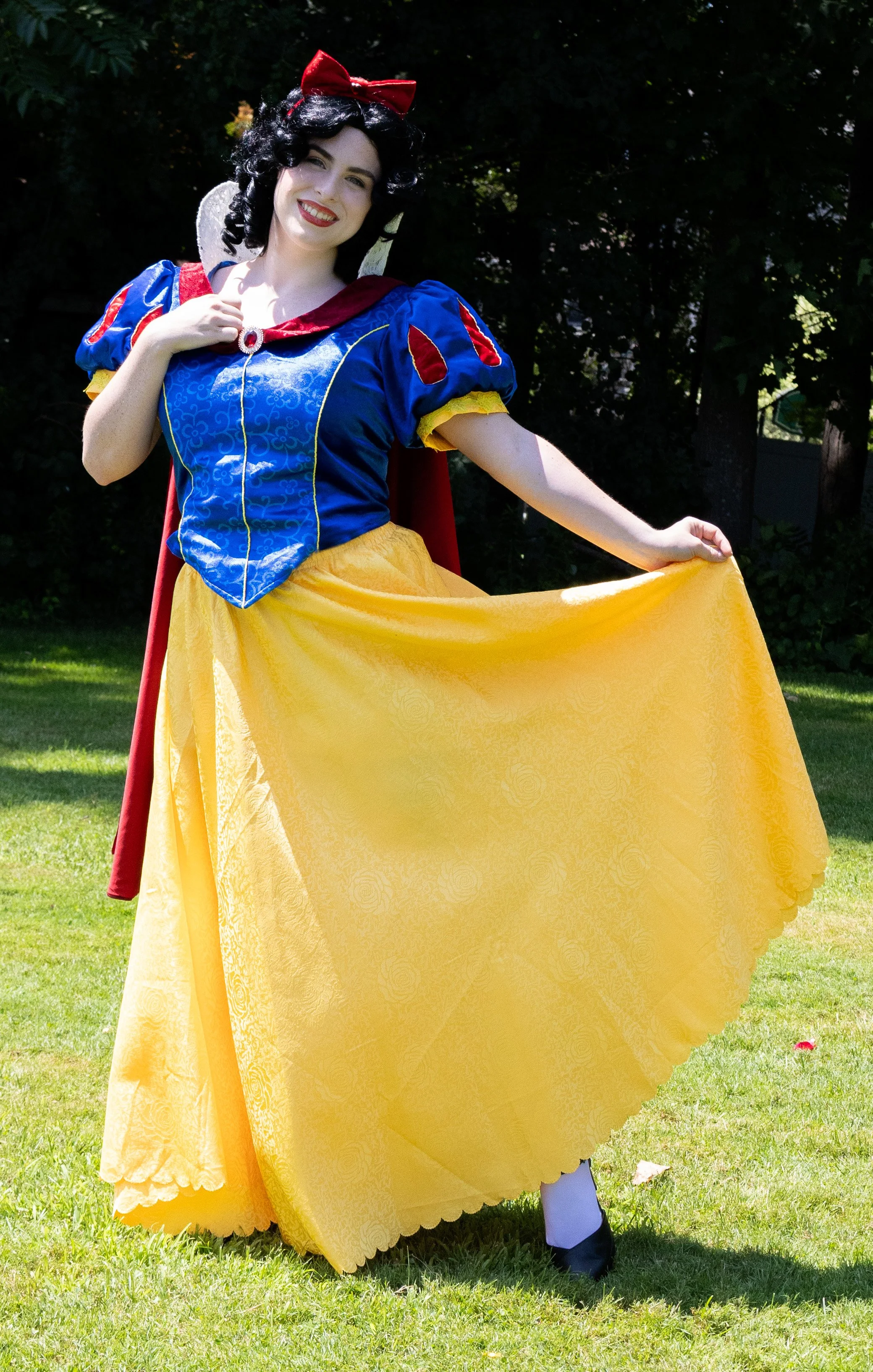 Snow White party princess standing outdoors on grass, holding up her yellow skirt with one hand, with a background of green trees and foliage.