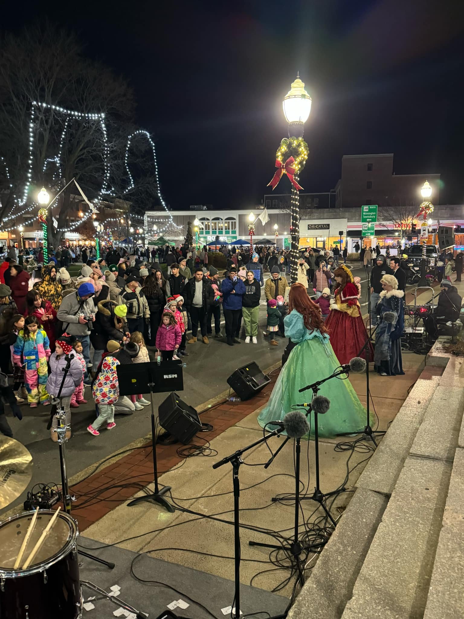 A Christmas-themed outdoor event with people gathered to watch party princess performers on stage. Christmas decorations, string lights, and wreaths are visible, with a lively crowd on a city street at night.