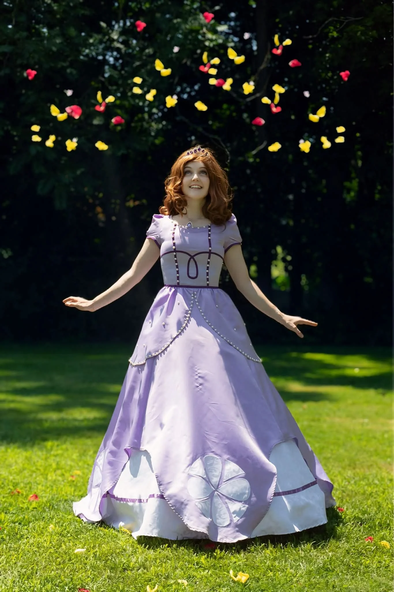 First Princess party character wearing a lavender ball gown with floral embellishments and a tiara, standing on a grassy lawn with trees in the background. 