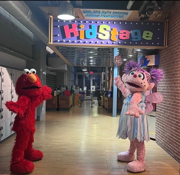 Furry Red Monster and Fairy in Training Party Characters at the Kids Stage entrance in Boston's Children's Museum with lockers and seating in the background.