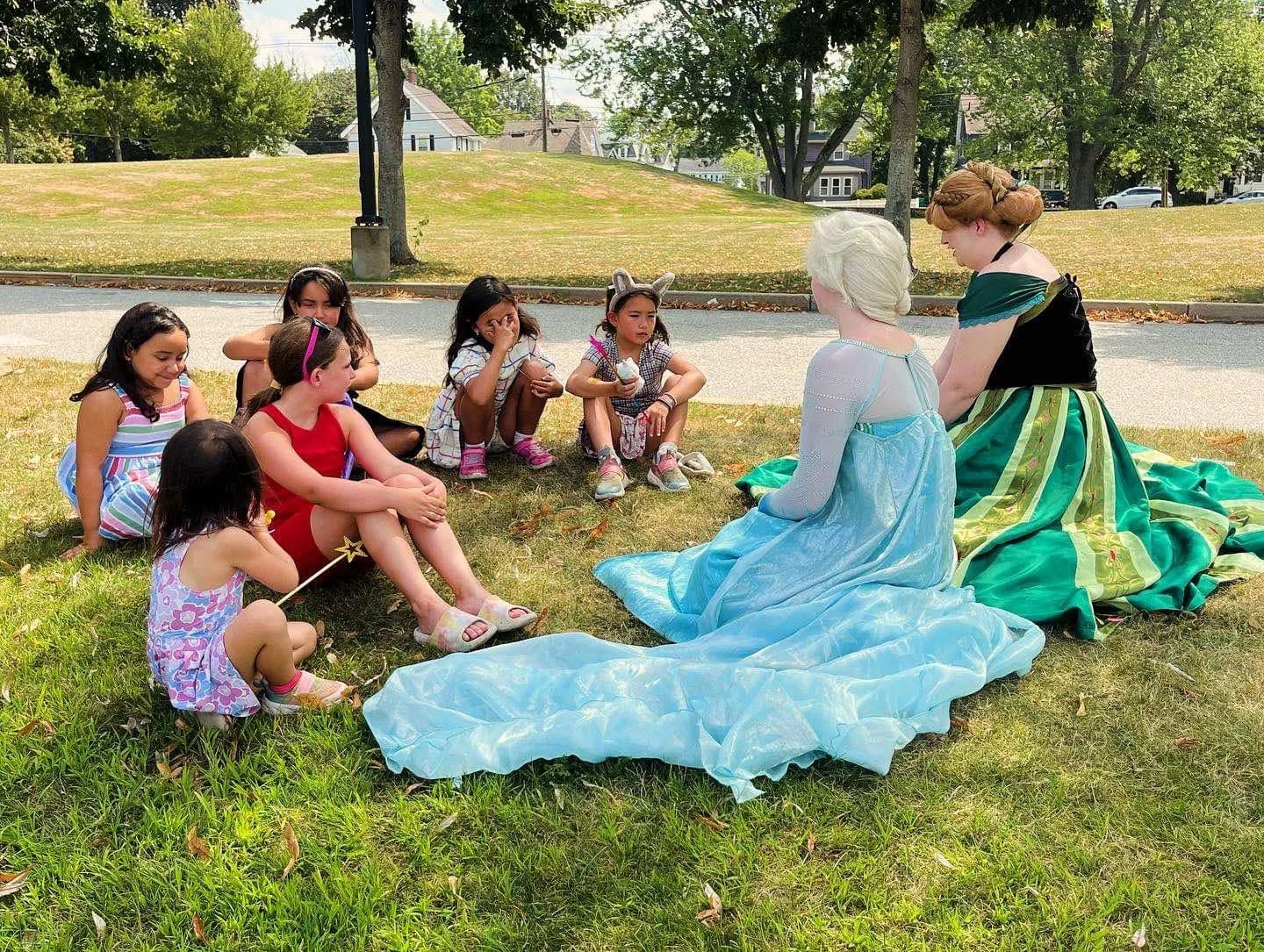 Children and Snow Sisters party princess characters sitting on grass in a park, engaging in storytelling or a conversation.