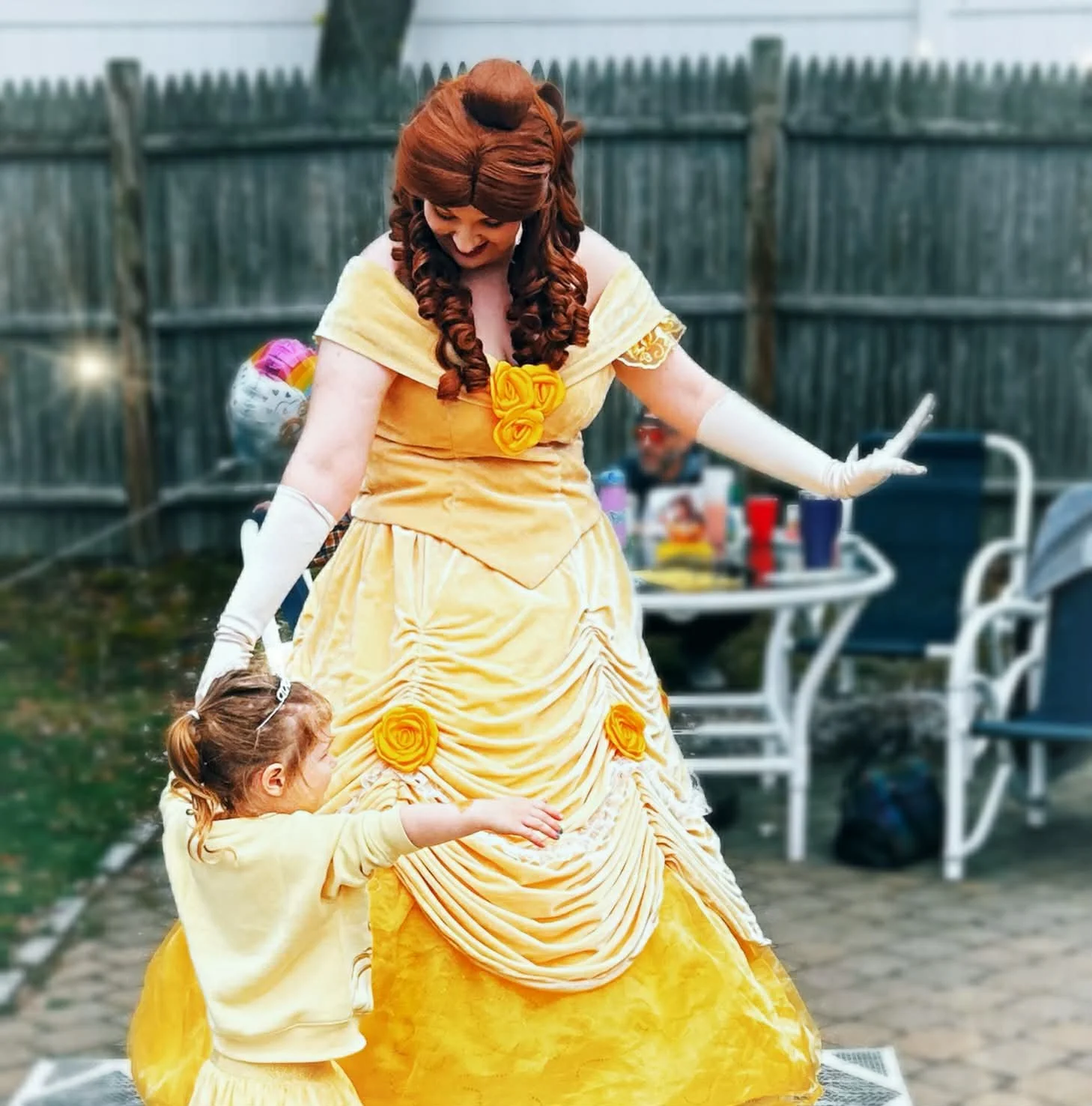 Princess Beauty party princess interacting with a young girl in a yellow dress resembling her gown, outside in a backyard with a wooden fence and patio furniture.