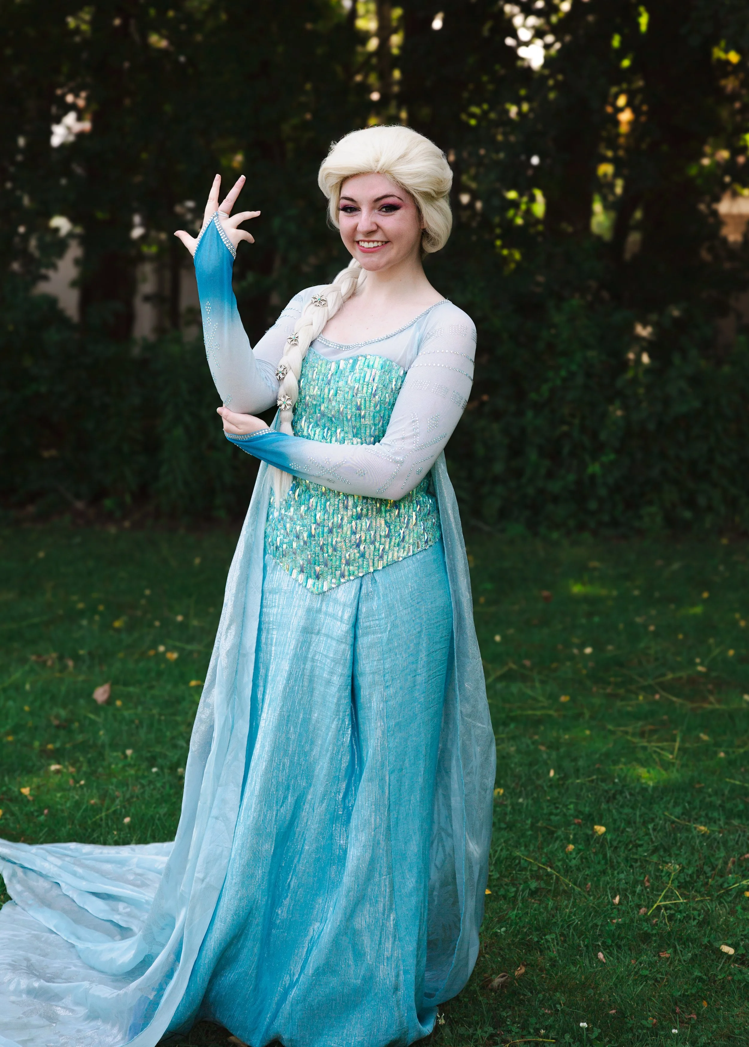 Snow Queen party princess wearing a light blue gown with a sheer cape and long illusion sleeves, standing outdoors on grass with trees in the background.